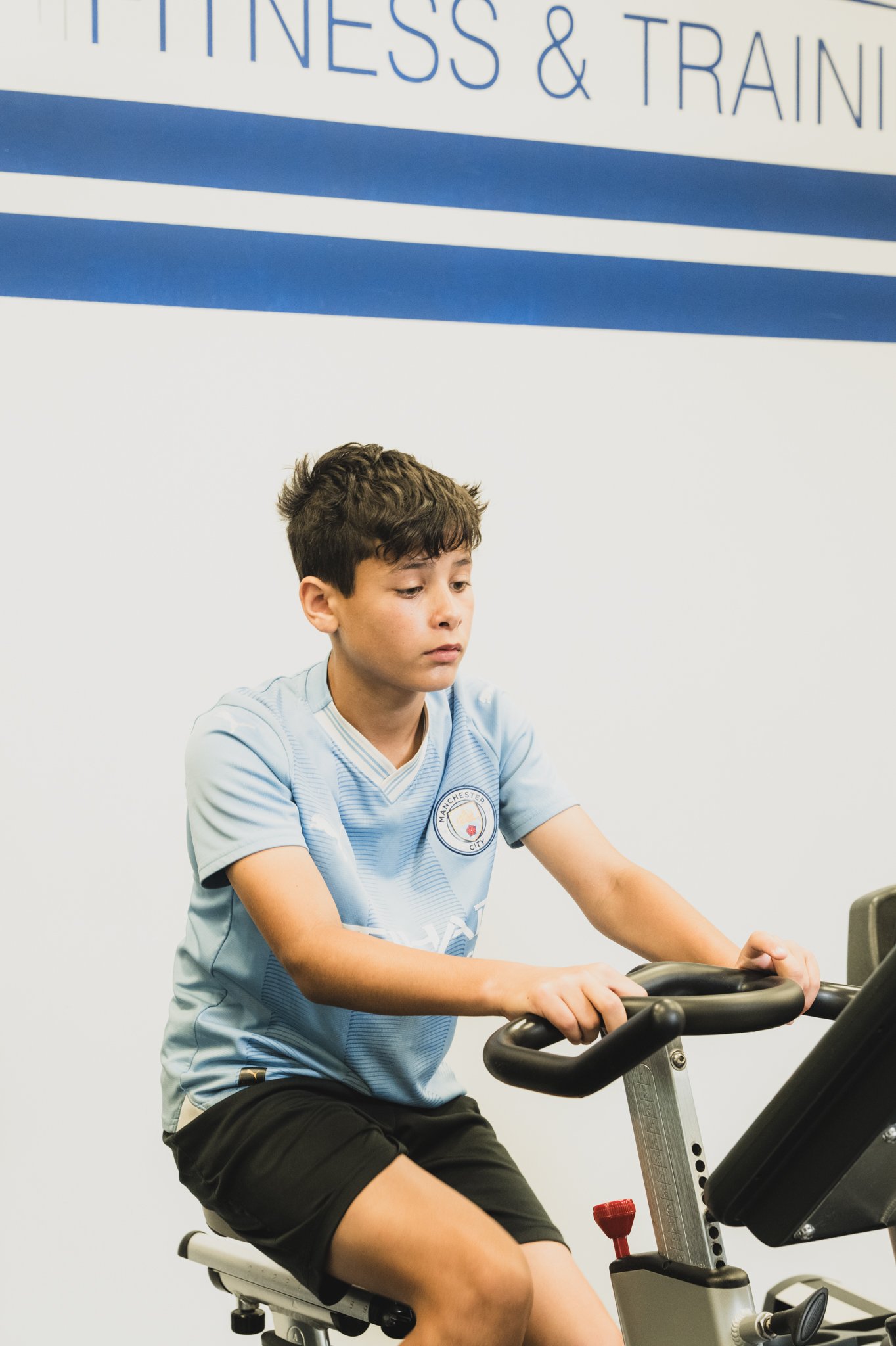 Young male with brown hair wearing a light blue Manchester City sports jersey riding a stationary exercise bike in a fitness center.