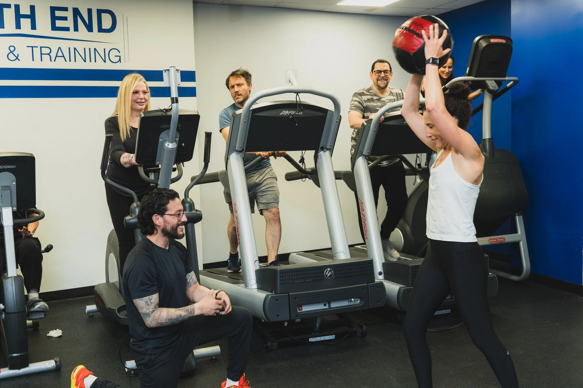 A woman lifting a medicine ball on a treadmill with two men and another woman observing in a gym.
