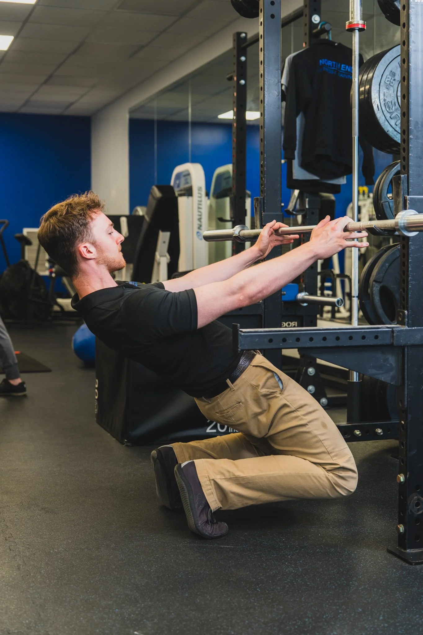 A young man in khaki pants and a black shirt is performing a kneeling back squat with a barbell in a gym. He is in a squat rack, gripping the barbell, with weight plates on its sides. The gym has a mirror, exercise equipment, and a blue wall in the background.