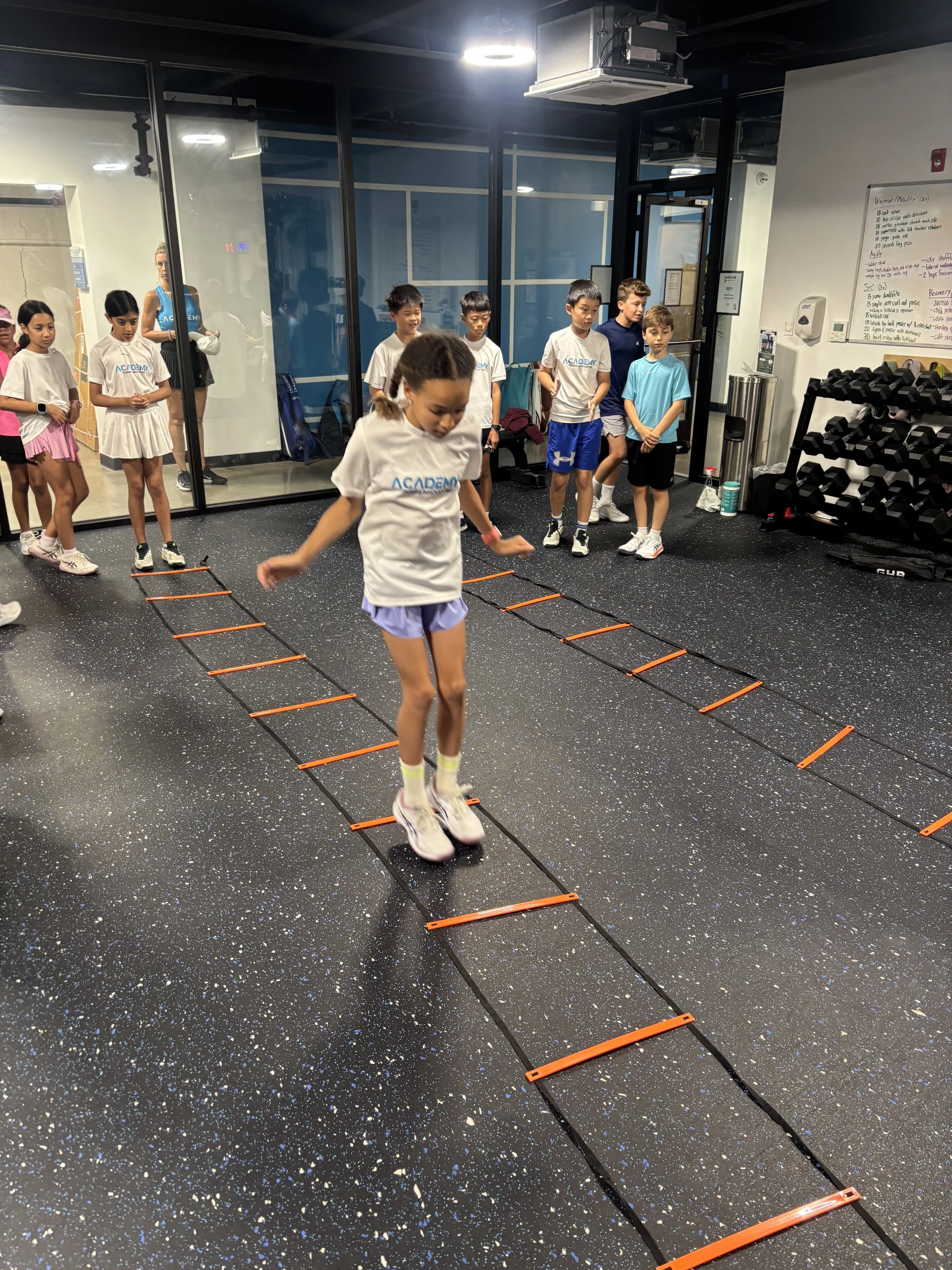 A group of children in athletic clothing participating in a drill involving agility ladders in a gym room with a speckled floor, a whiteboard, and equipment.