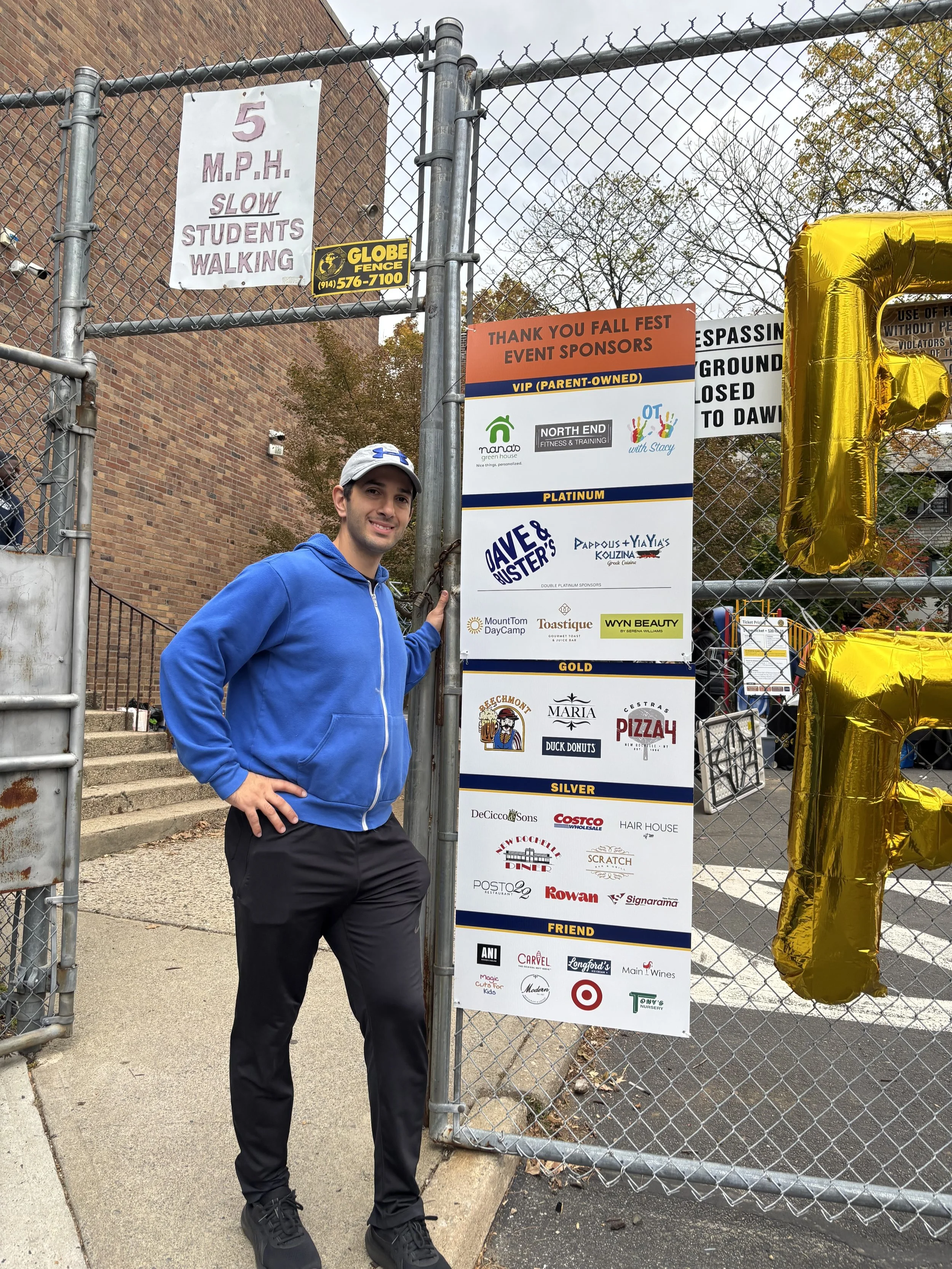 A man in a blue hoodie and black pants standing next to a fence with event sponsor signs. The signs thank sponsors for a fall festival, display best sponsor levels, and list various local businesses. A large gold balloon shaped like the letter R is partially visible on the right.