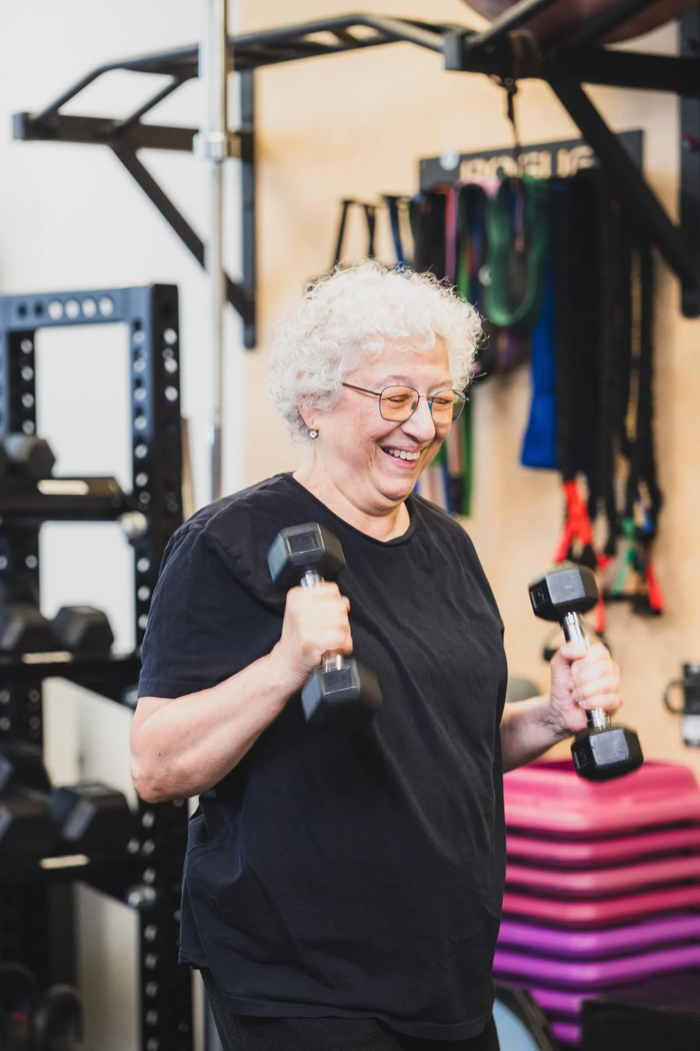A senior with curly white hair and glasses smiling while lifting black dumbbells in a gym. Shelves with colorful resistance bands and workout equipment are visible in the background.