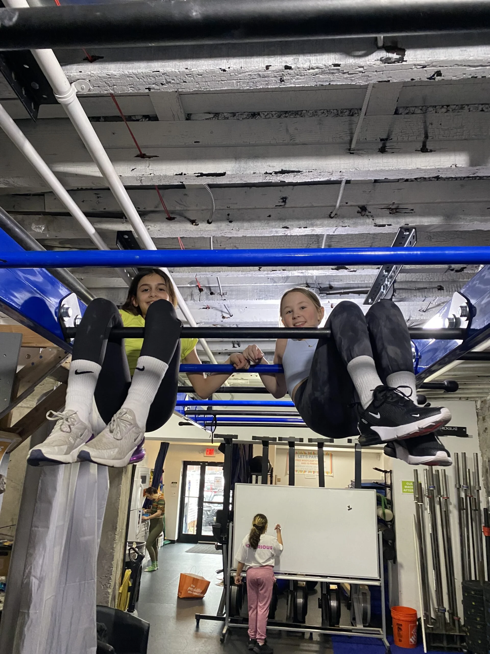Two children are sitting on an exercise machine, smiling and holding onto the bars. The photo is taken from below, showing the ceiling of a gym with wooden beams and pipes. In the background, a woman is writing on a whiteboard, and another person is near a door.