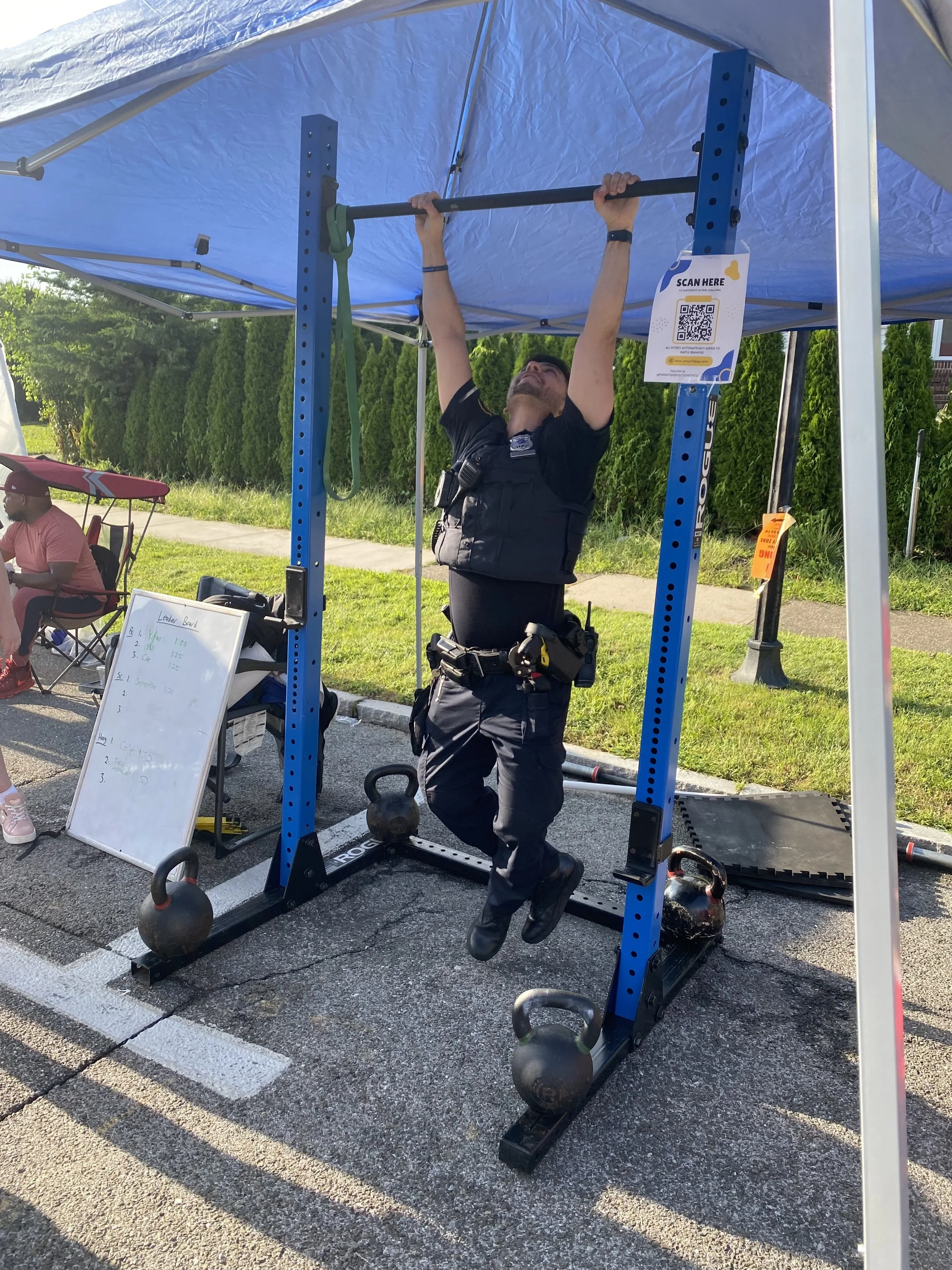 A man in a police uniform is inside a small outdoor gym tent, performing a pull-up exercise on a black bar. There are kettlebells on the ground, a whiteboard with notes, and a sign with a QR code attached to the tent. In the background, another person is seated near a folding chair, and green bushes with trees line the perimeter of the outdoor space.