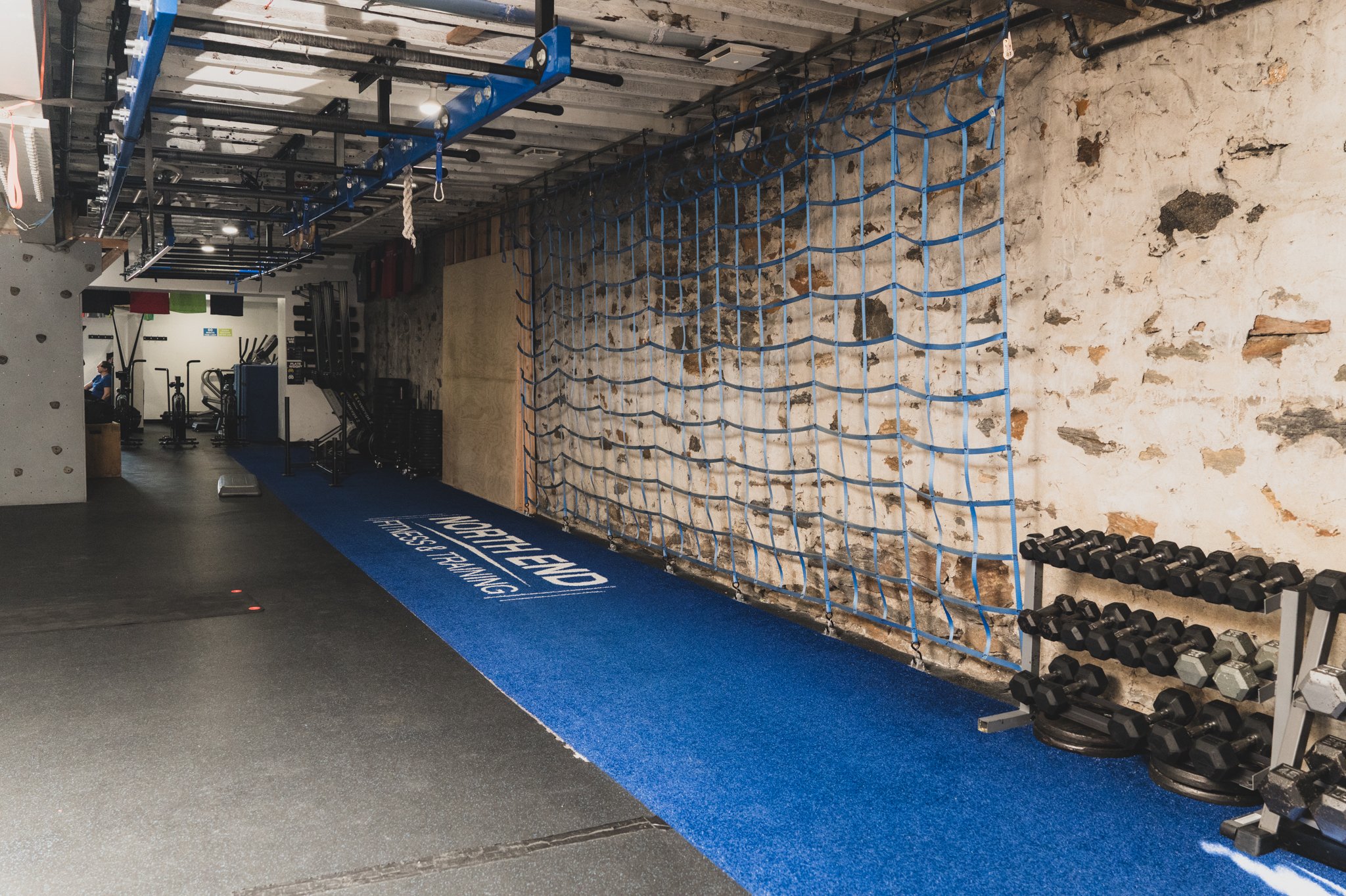 Inside a gym with workout equipment, a blue storage rack with resistance bands hanging, a wall with exposed brick, a blue mat with white lettering, a row of dumbbells, and a person working out in the background.