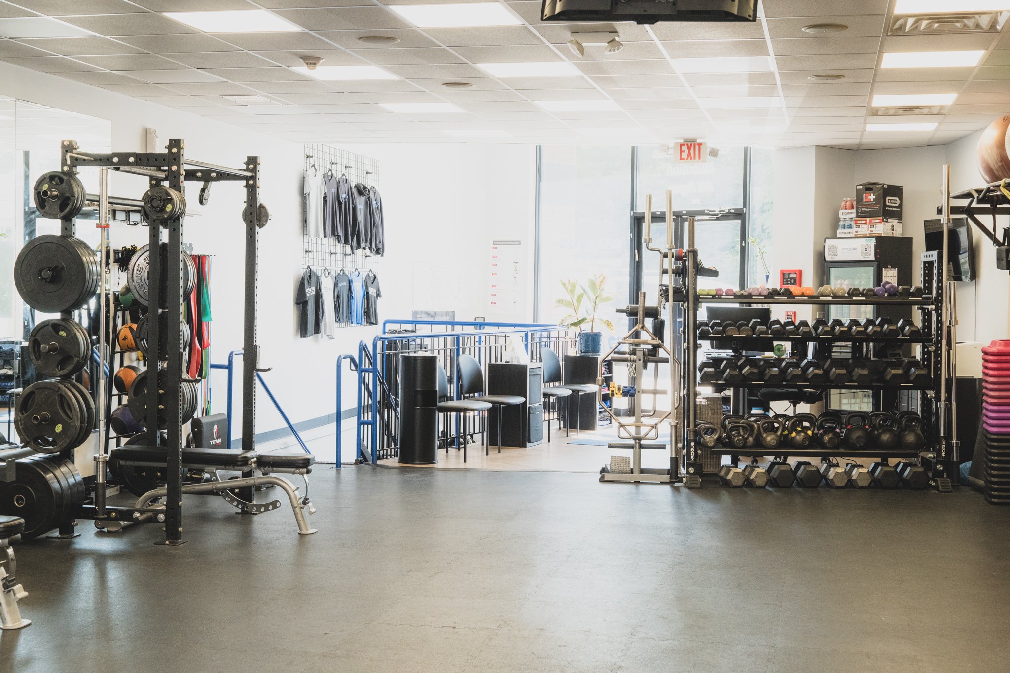 Interior of a gym with weightlifting equipment, like barbells, dumbbells, and medicine balls, along with racks of clothing, chairs, and an exit sign near the door.