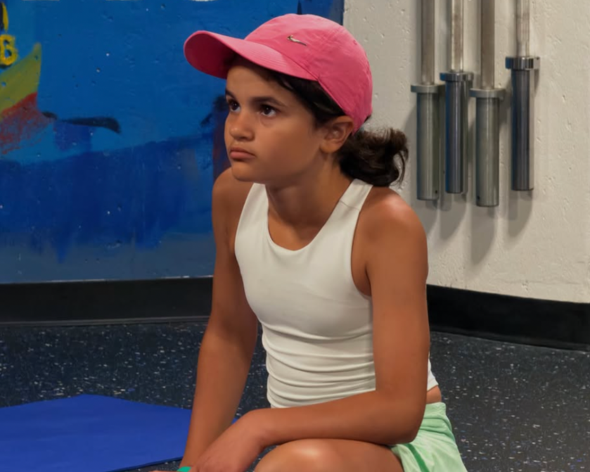 A young girl with dark hair, wearing a pink cap, white tank top, and light green shorts, sitting on the floor in a gym or indoor activity center, looking pensively to the side.
