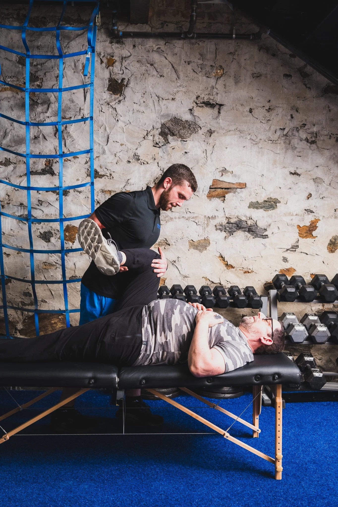 Two men working out in a gym with a trainer assisting one of them. The man lying on the workout bench is holding his hand to his chest, wearing glasses and a tiger stripe shirt. The trainer is standing beside him, supporting his knee, wearing a black shirt and blue shorts. The gym has a rustic brick wall background, with dumbbells on racks and a blue rope climbing net on the side.