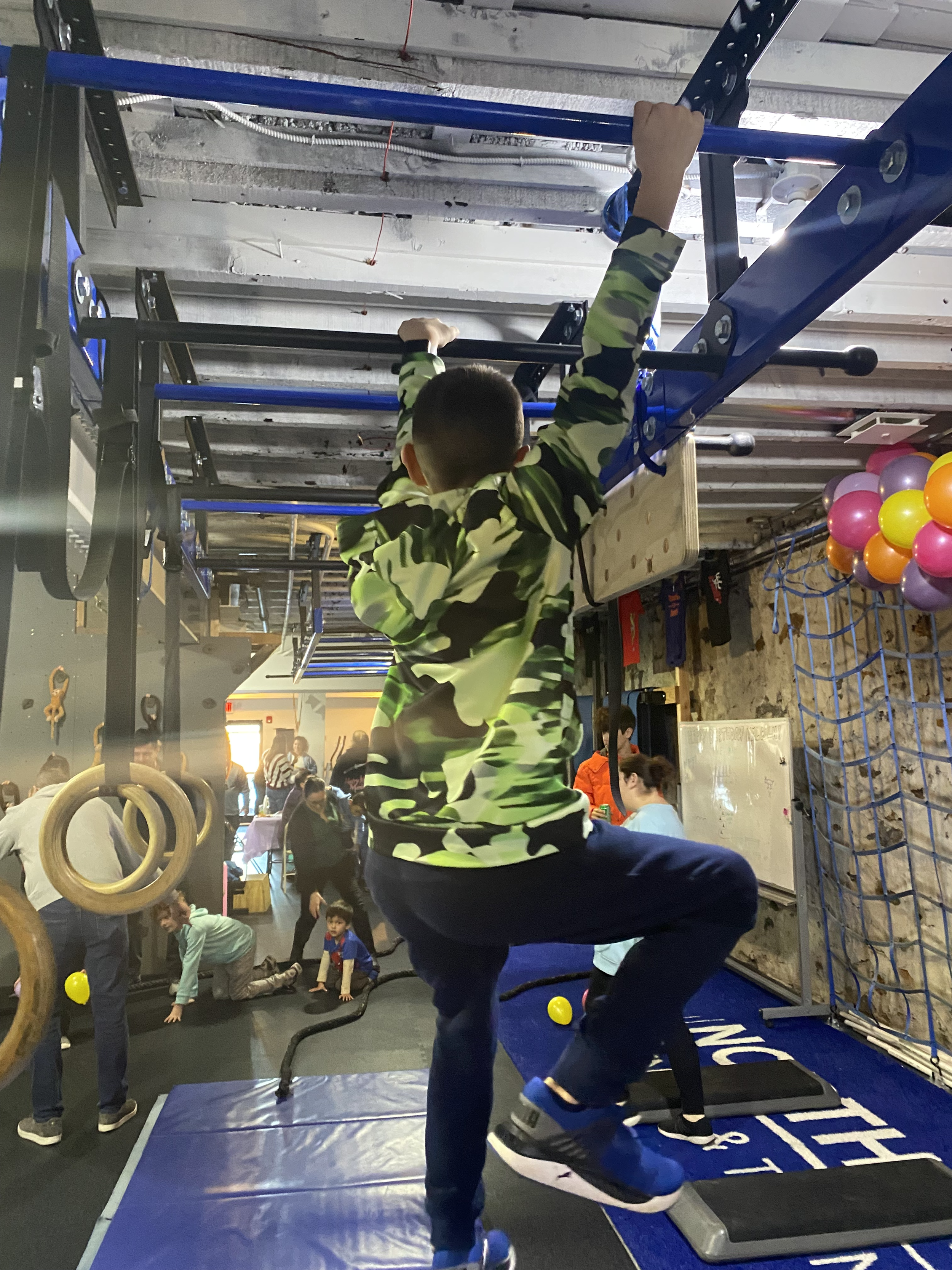 A young boy in camo sweatshirt is climbing a wall in an indoor gym. Other children and adults are visible in the background, along with gym equipment, balloons, and a netted area.