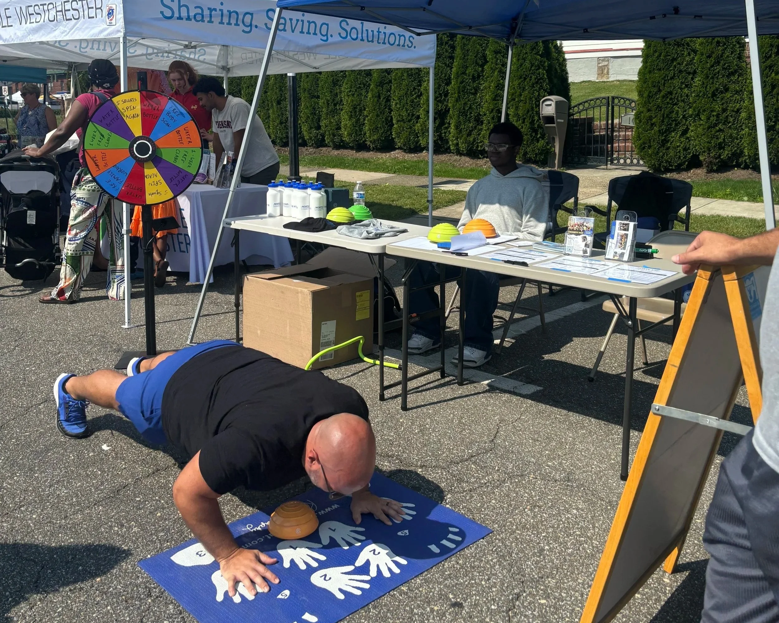 A man doing a plank exercise on a blue yoga mat outdoors at a community event booth with a table and a prize wheel. Other people are seen in the background under tents.