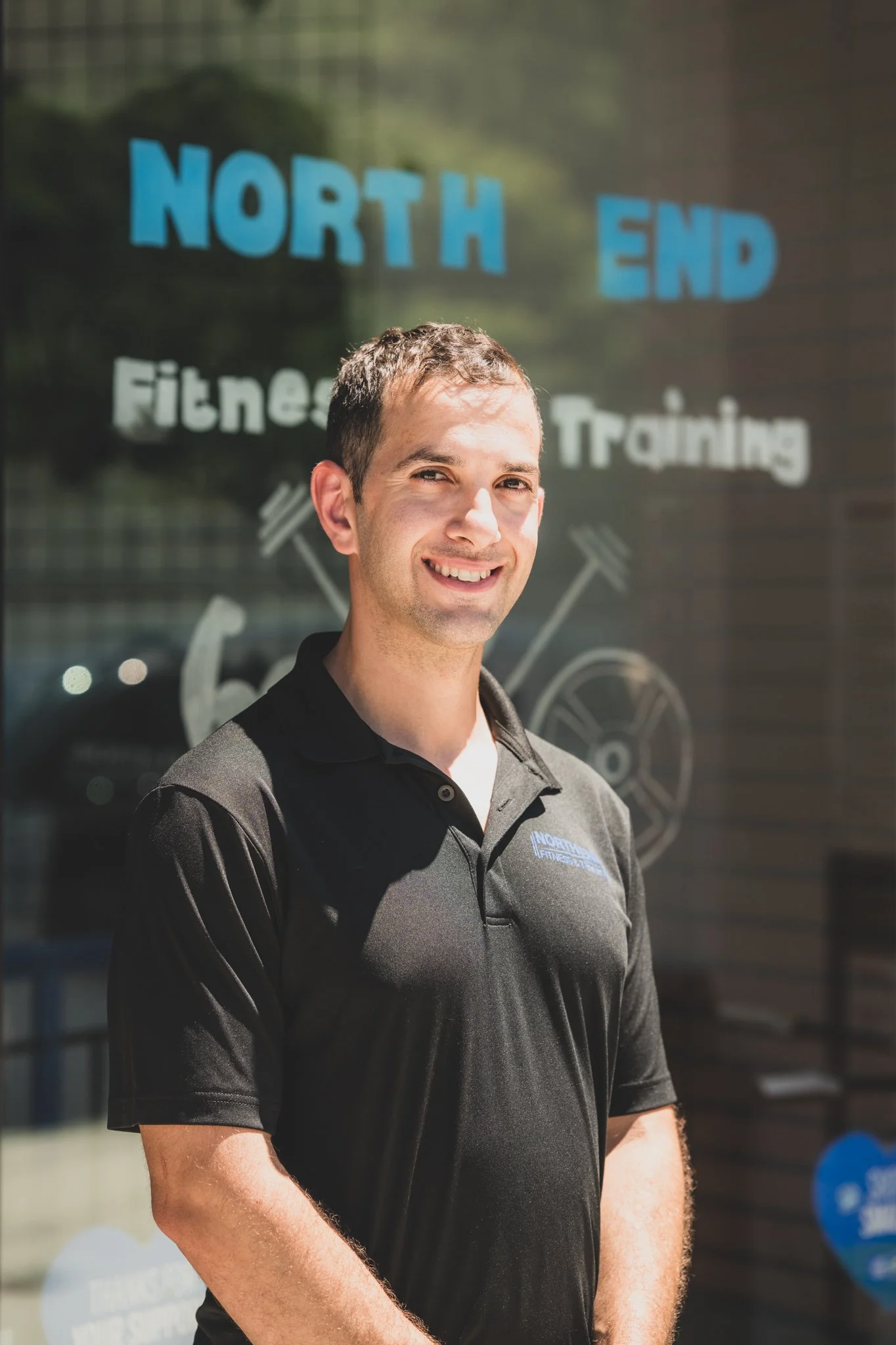 A young man smiling outdoors in front of a glass window with writing about fitness and training, wearing a black polo shirt with a logo.