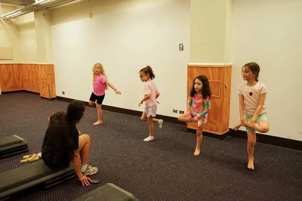 Five young girls practicing a dance or exercise routine in a room with wood-paneled walls and carpeted floor, while a woman sitting on a bench observes.