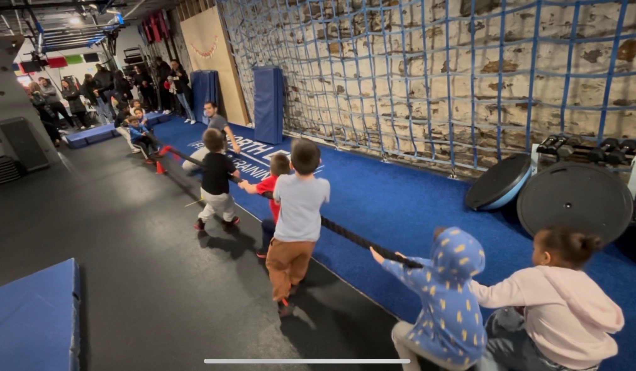 Children and adults participating in a tug-of-war game at an indoor event space. The children are pulling a rope while the adults watch nearby. The background features a rock climbing wall and people socializing.