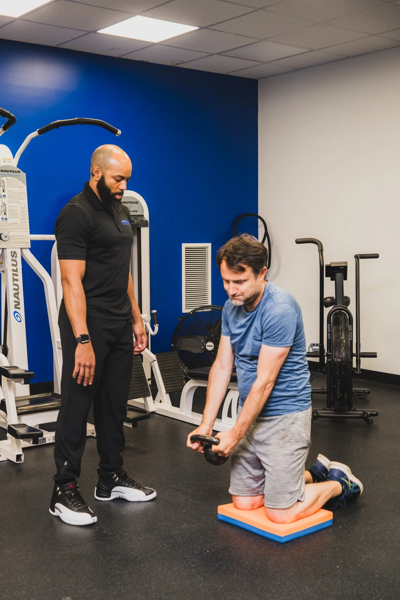 A man is kneeling on a cushioned exercise pad while holding a kettlebell with both hands in a gym. A personal trainer stands nearby, observing and providing guidance. The gym equipment and a fan are visible in the background.