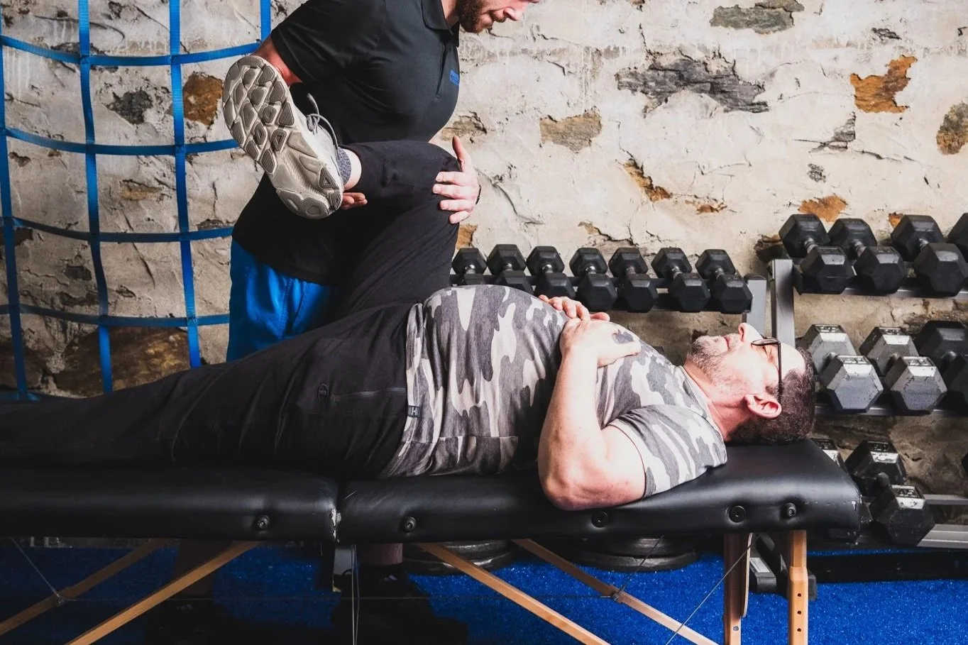 A man lying on a therapy table receiving assistance for a leg injury, with a trainer supporting his leg, in a gym with dumbbells on racks and a stone wall background.