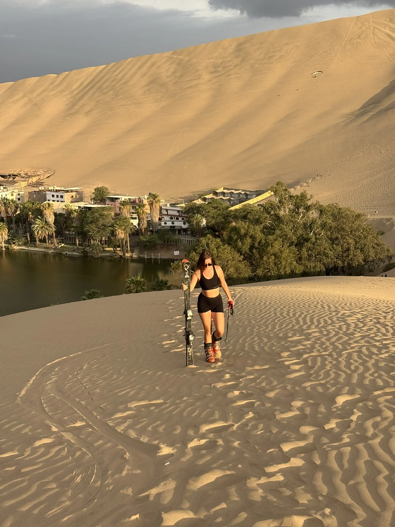 Una mujer con ropa deportiva y botas de esquí subiendo una duna de arena, con un paisaje de dunas, árboles y casas en el fondo, y un cielo nublado.