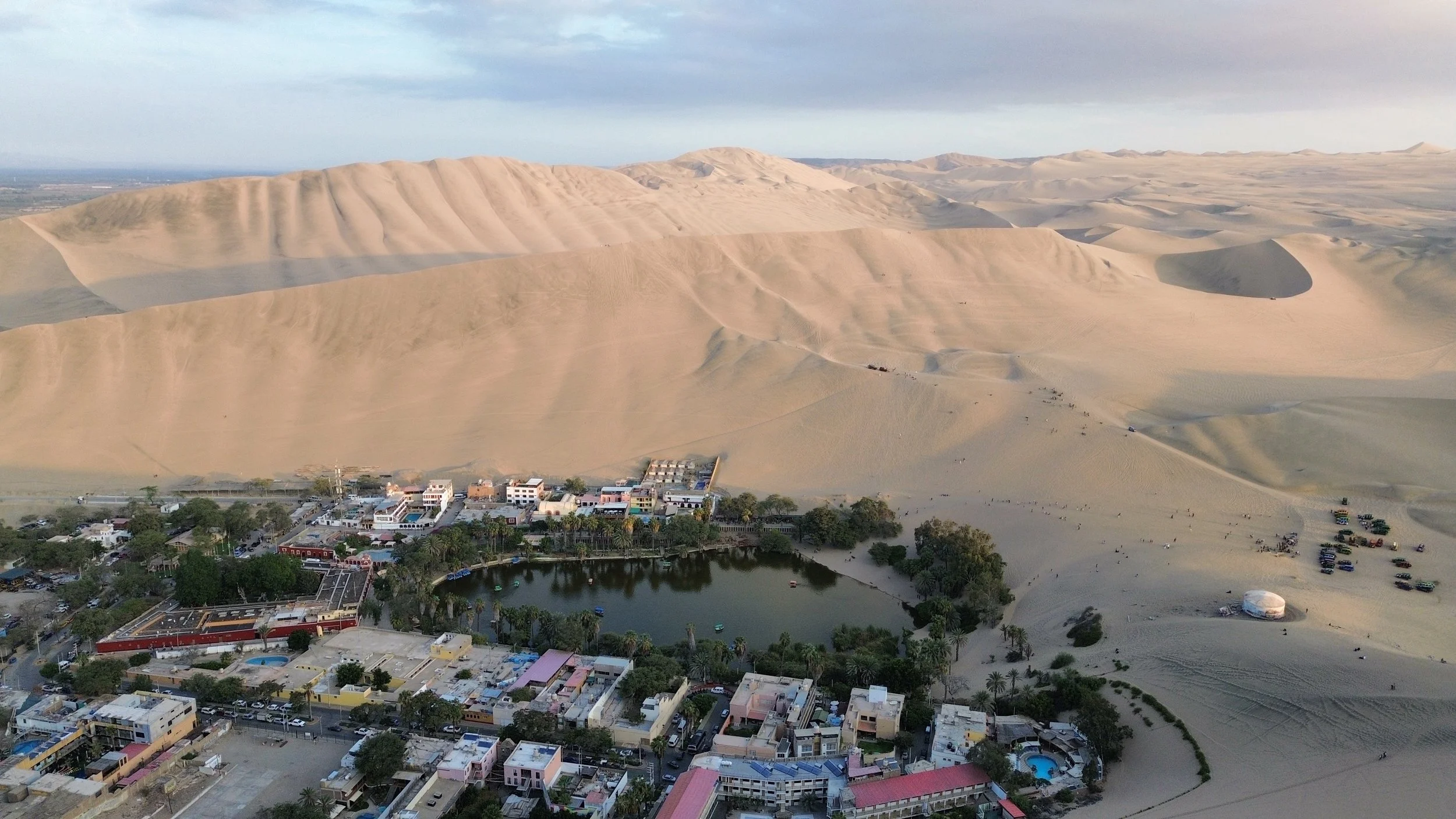 Una vista aérea de las dunas de arena en un paisaje desértico con un pueblo pequeño y un cuerpo de agua en primer plano.