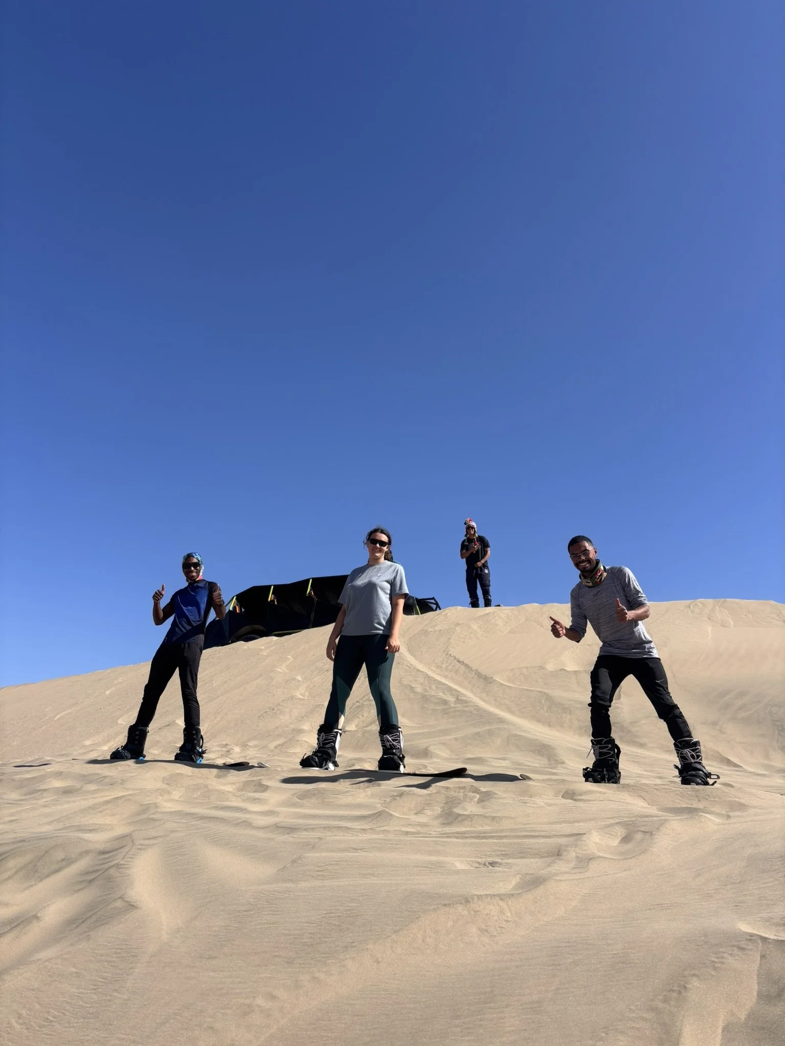 Cuatro personas en sandboard en un desierto de dunas, sonriendo y posando para la cámara bajo un cielo azul claro.