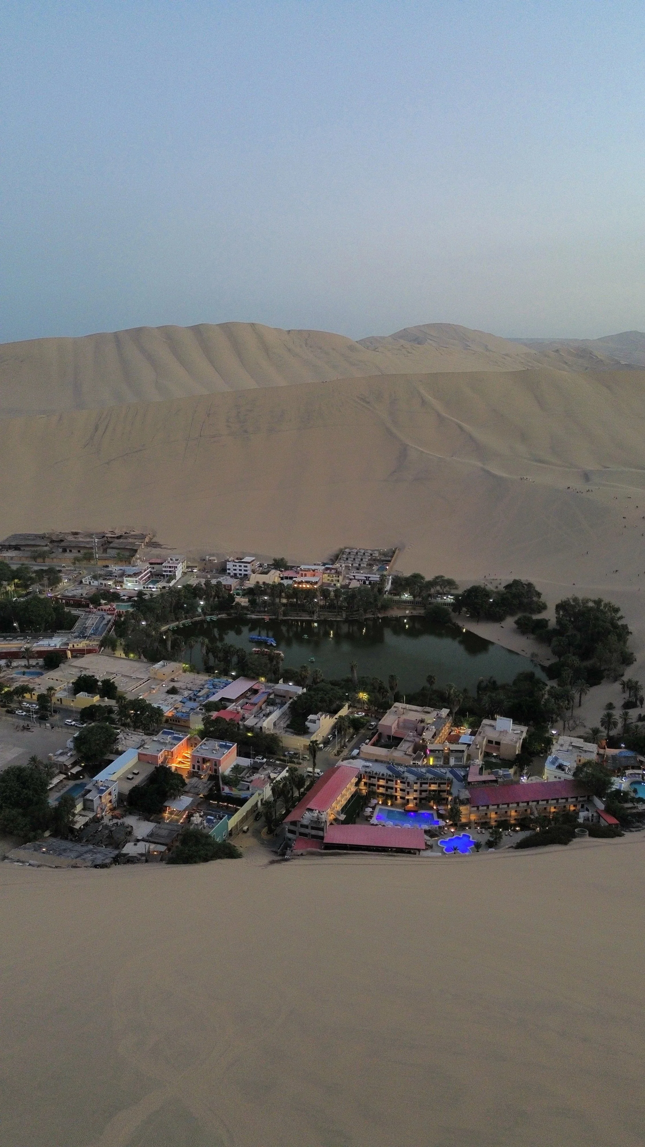Ciudad con un oasis, edificios y una piscina, rodeada de arena y un desierto en el fondo, al atardecer