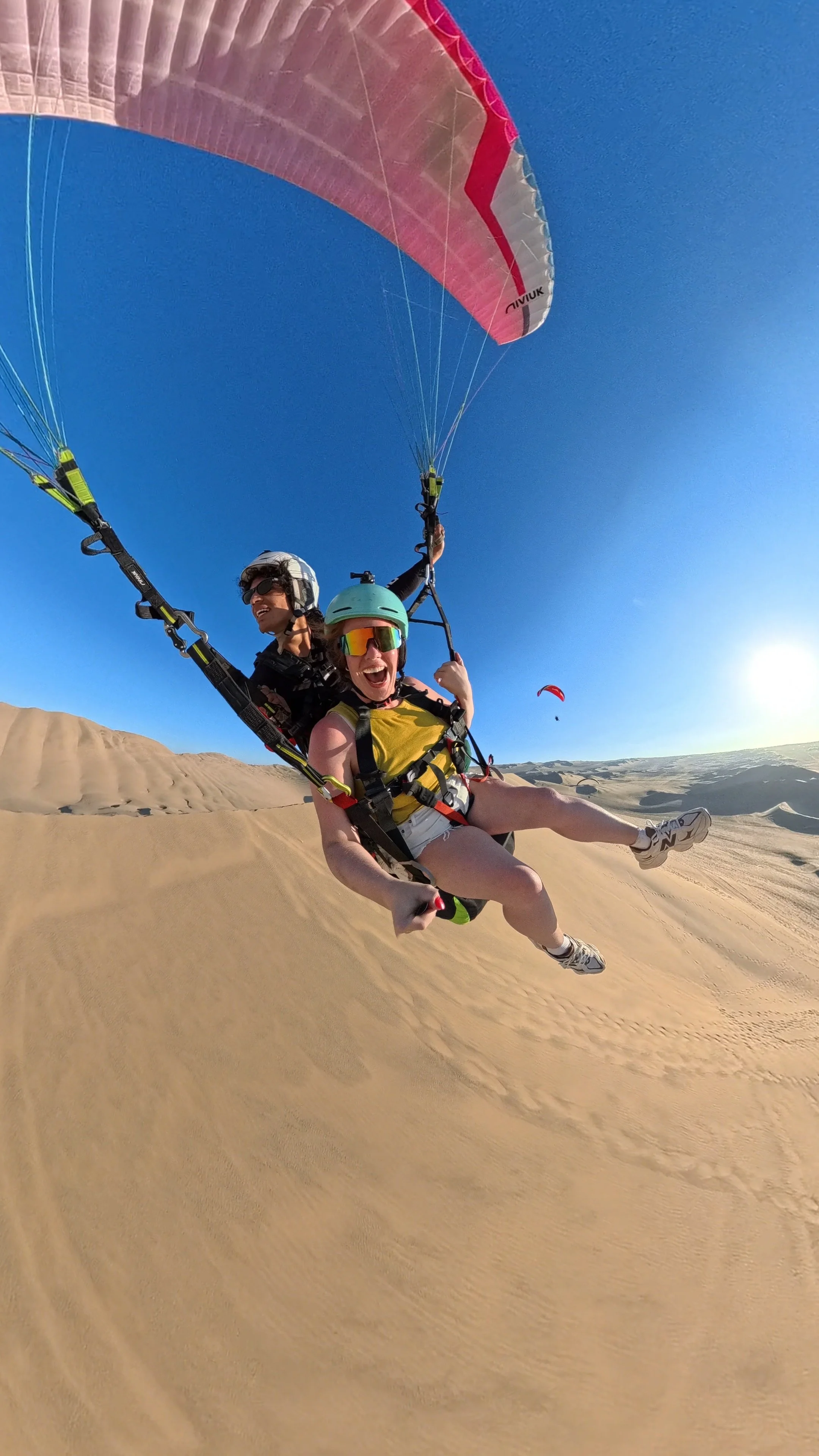 Dos personas practicando parapente en un desierto, con una mujer en primer plano (pasajera) sonriendo y usando casco y gafas de sol, y otra persona (el piloto) en el fondo también con casco y gafas, con un cielo azul despejado.
