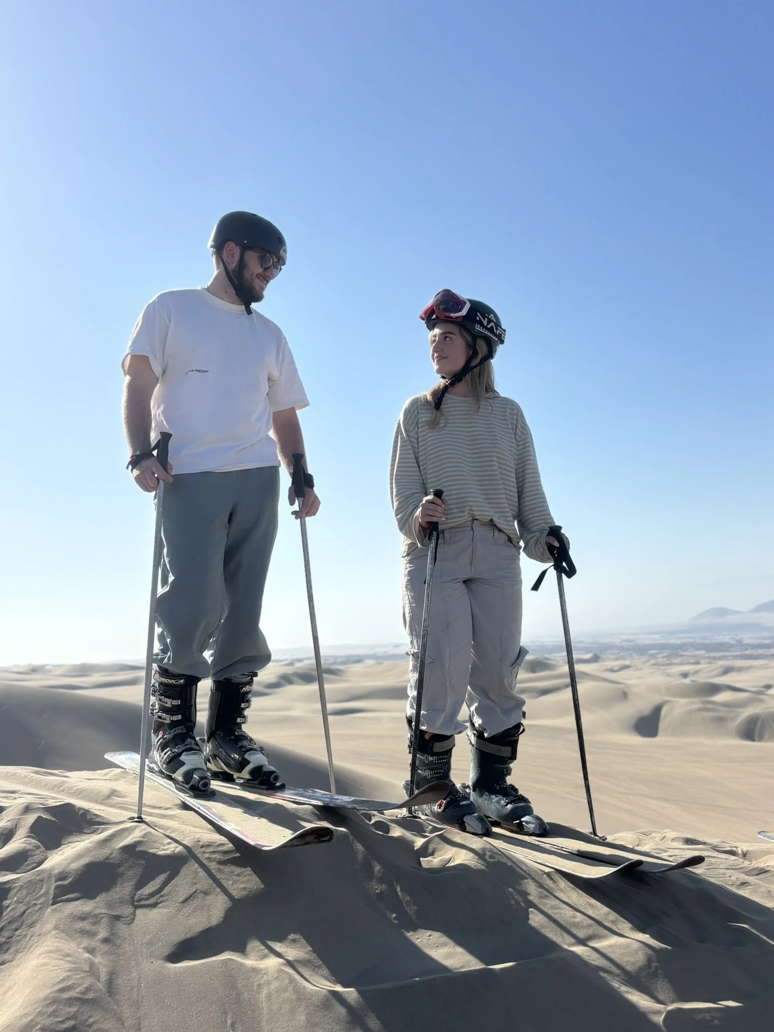 Dos personas con cascos y esquís en un paisaje de dunas de arena, disfrutando del día soleado.