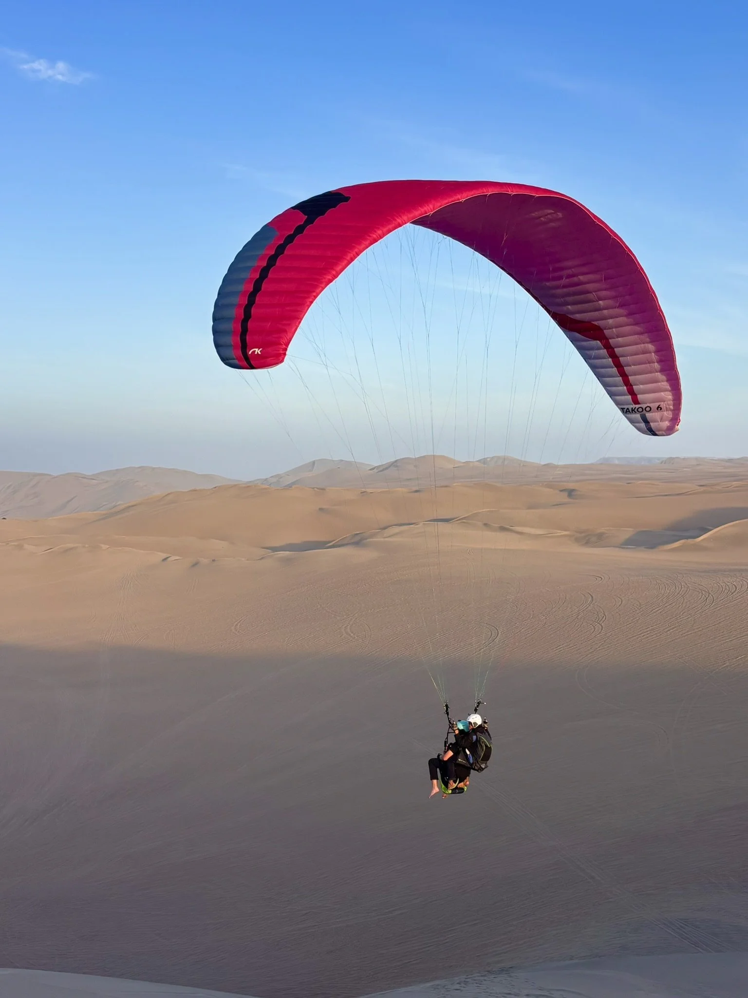 Persona practicando parapente tandem en un desierto con dunas, cielo despejado y escasos nubes.