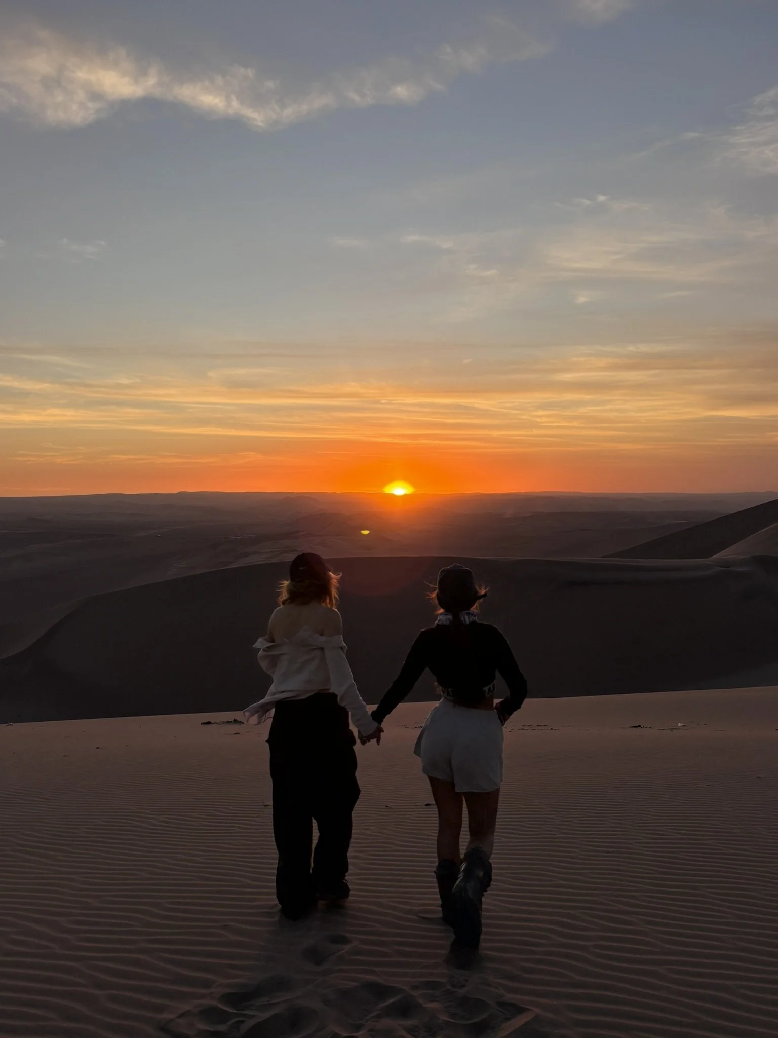 Dos personas caminando en dunas de arena durante un atardecer, con cielo despejado con algunas nubes.