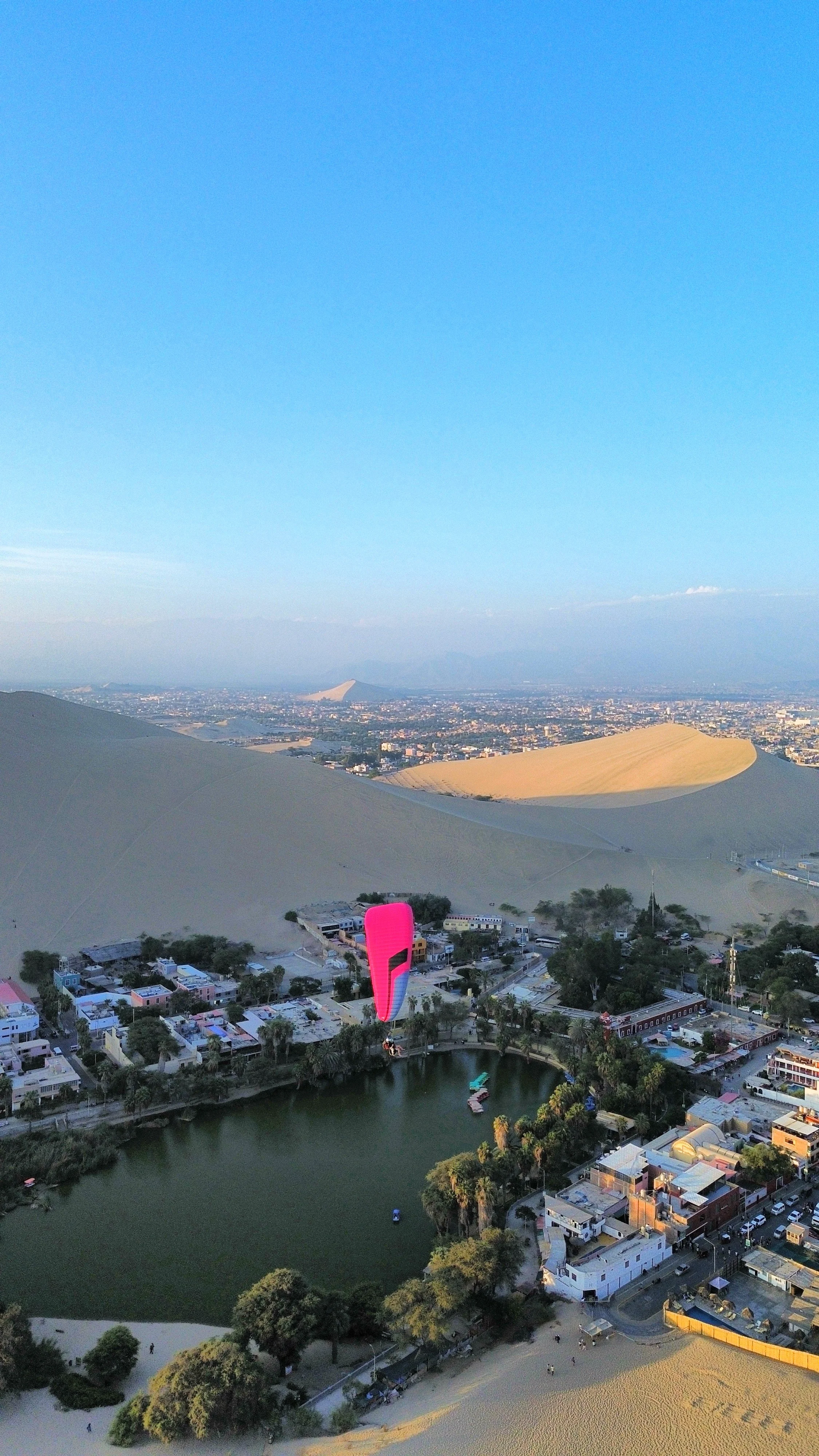 Ala desértica con dunas doradas y una ciudad en el fondo, un parapente rosado volando sobre un oasis y edificios alrededor. 