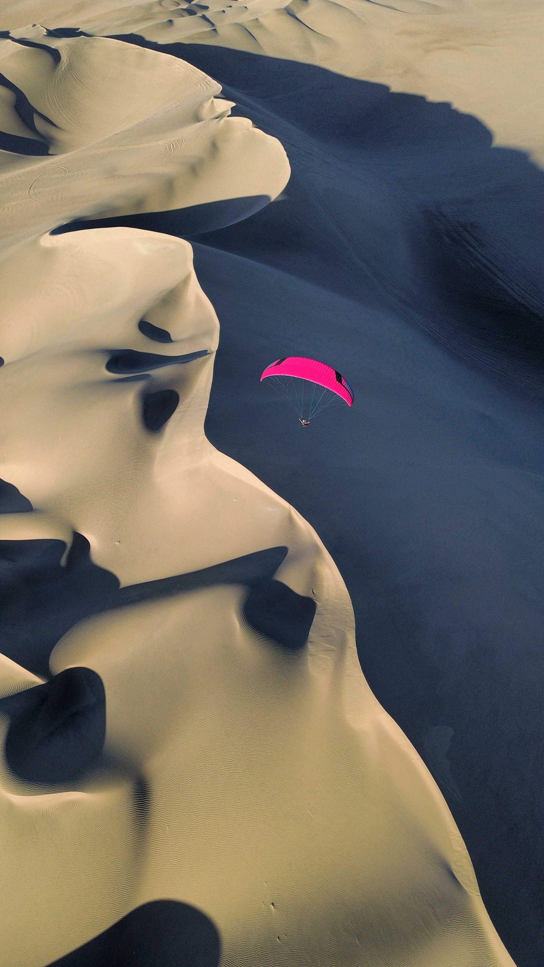 Una persona haciendo parapente sobre un paisaje de dunas de arena, con un parapente de color rosa.
