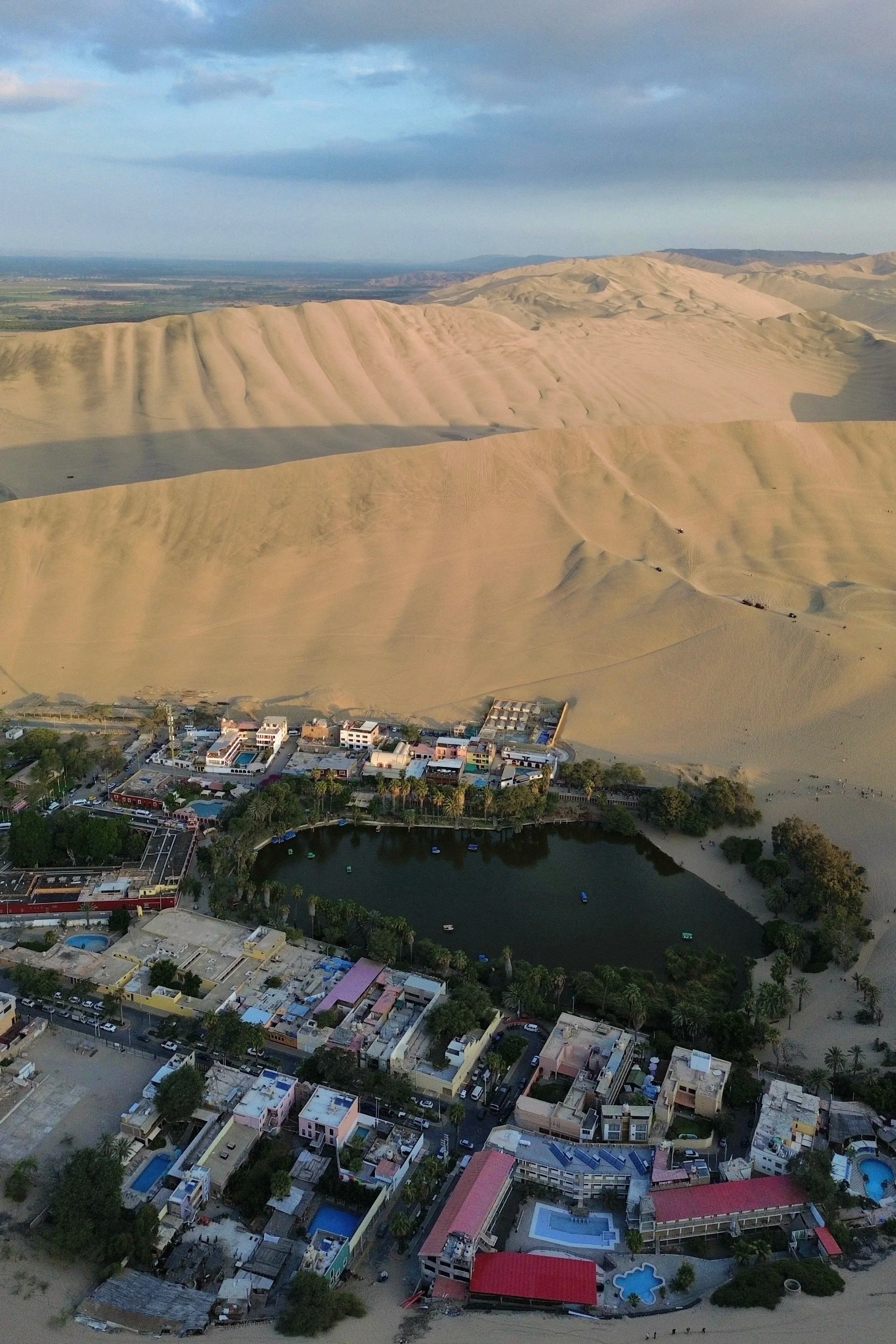 Vista aérea de un pueblo junto a un oasis, con un área de dunas de arena en el fondo y hoteles con piscinas.