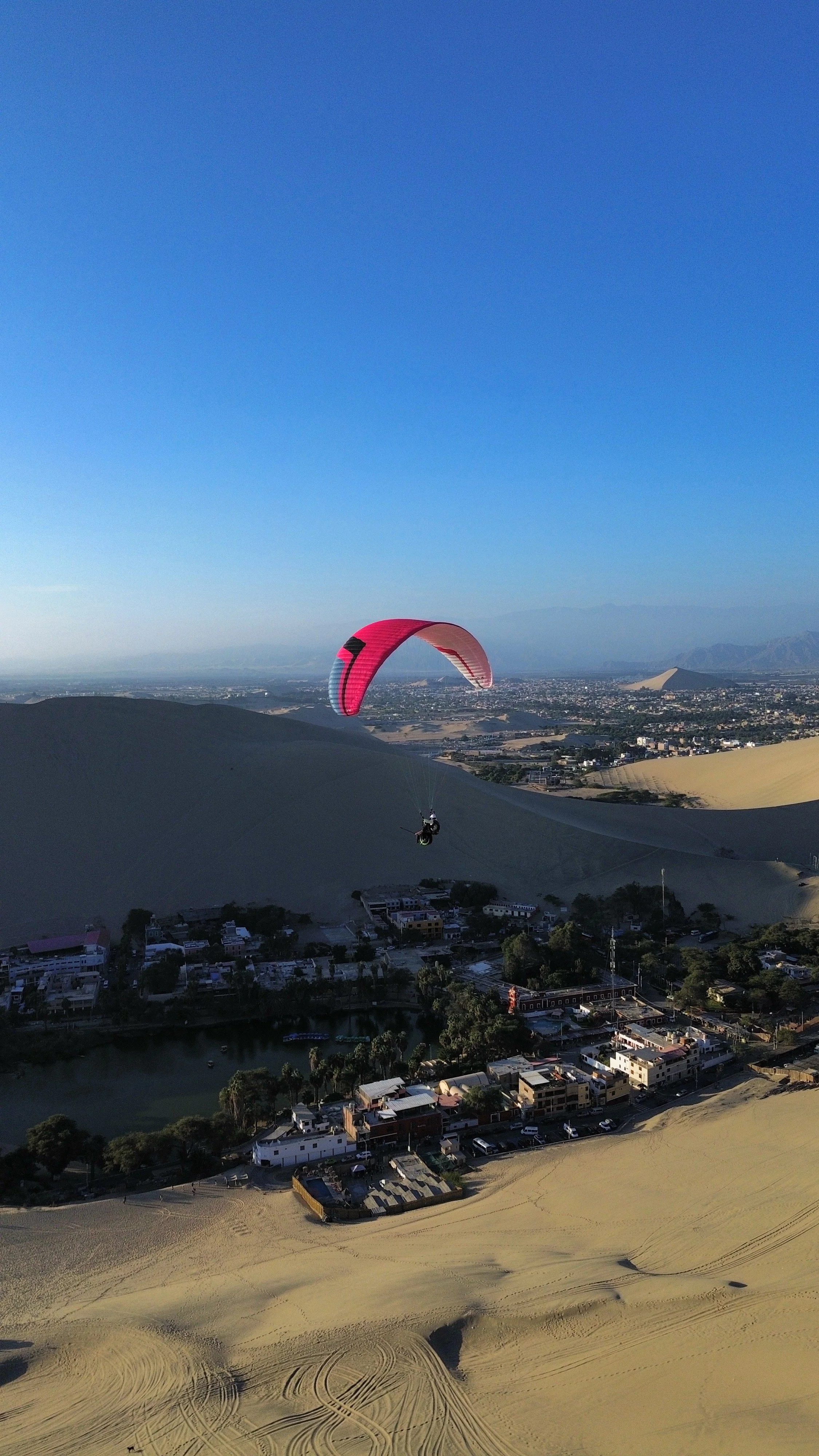 Persona realizando parapente sobre un paisaje de dunas y ciudad, con dunas al fondo y cielo despejado