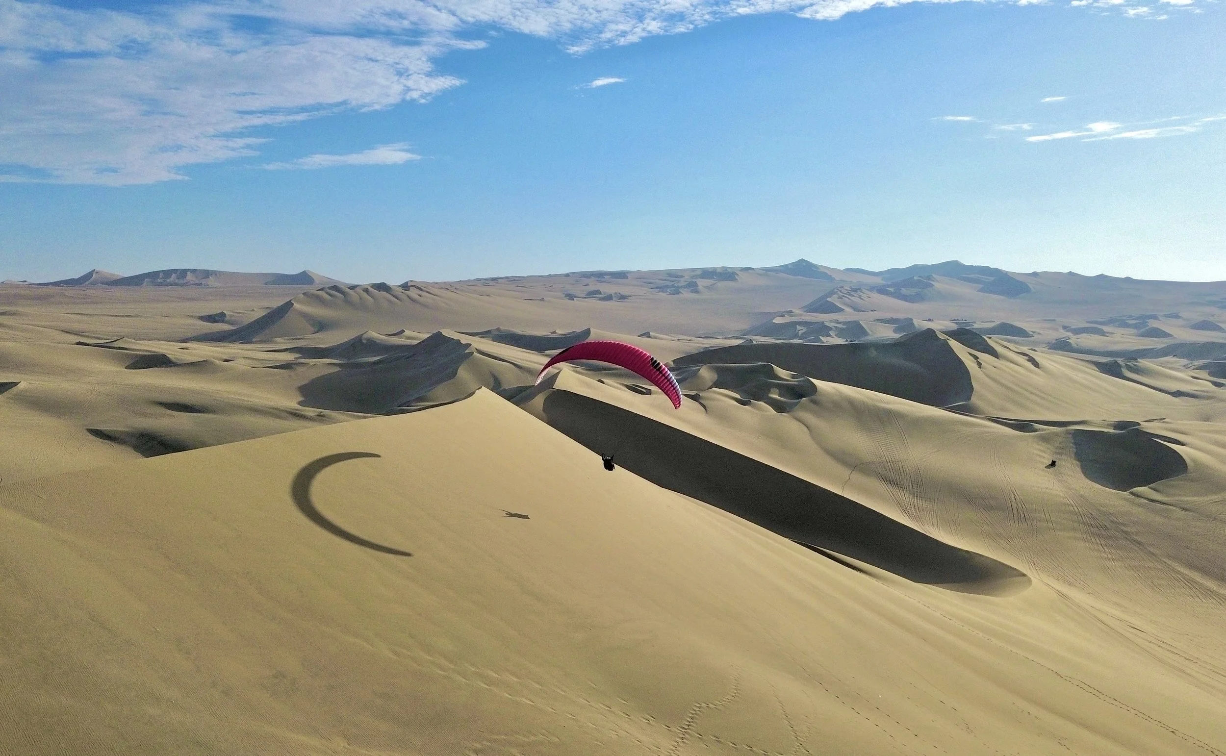 Un desierto con dunas de arena y un cielo despejado, con un parapente volando en el aire.