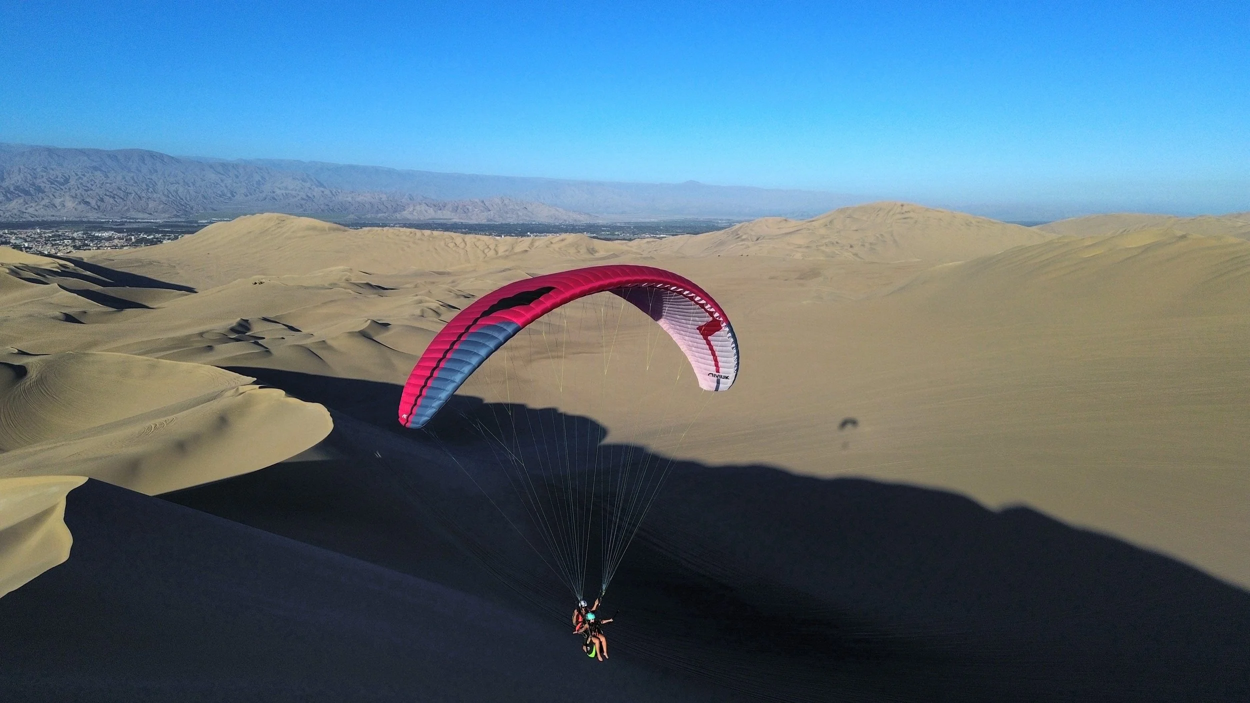 Piloto de parapente volando con un pasajero sobre dunas en un desierto con montañas al fondo y un cielo despejado.