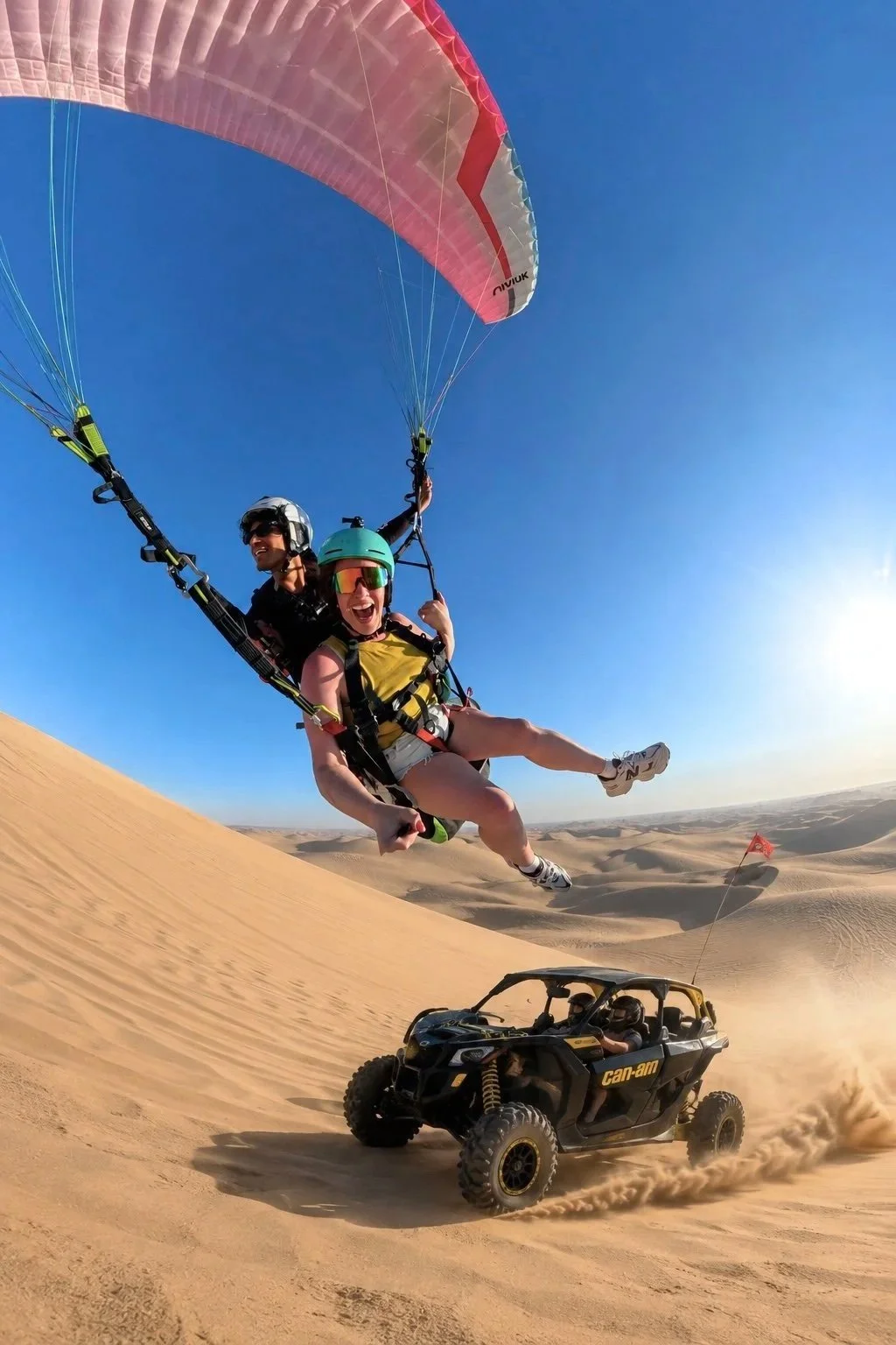 Dos personas haciendo parapente en un desierto, y un vehículo todoterreno en la arena.