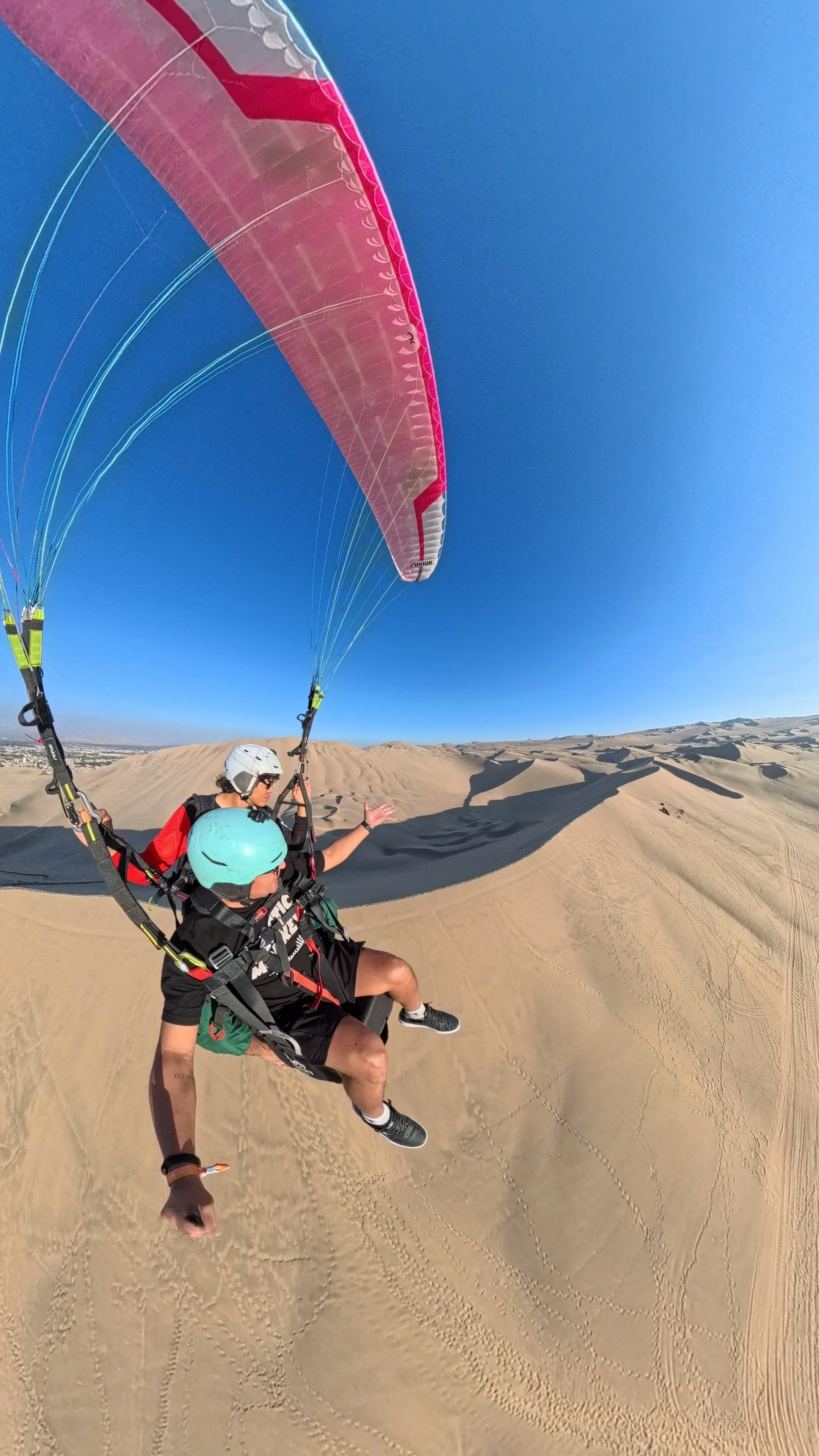 Dos personas con equipo de parapente sobre dunas de arena en un desierto, con casco y gafas, ambas con arnés, volando juntos en el aire.