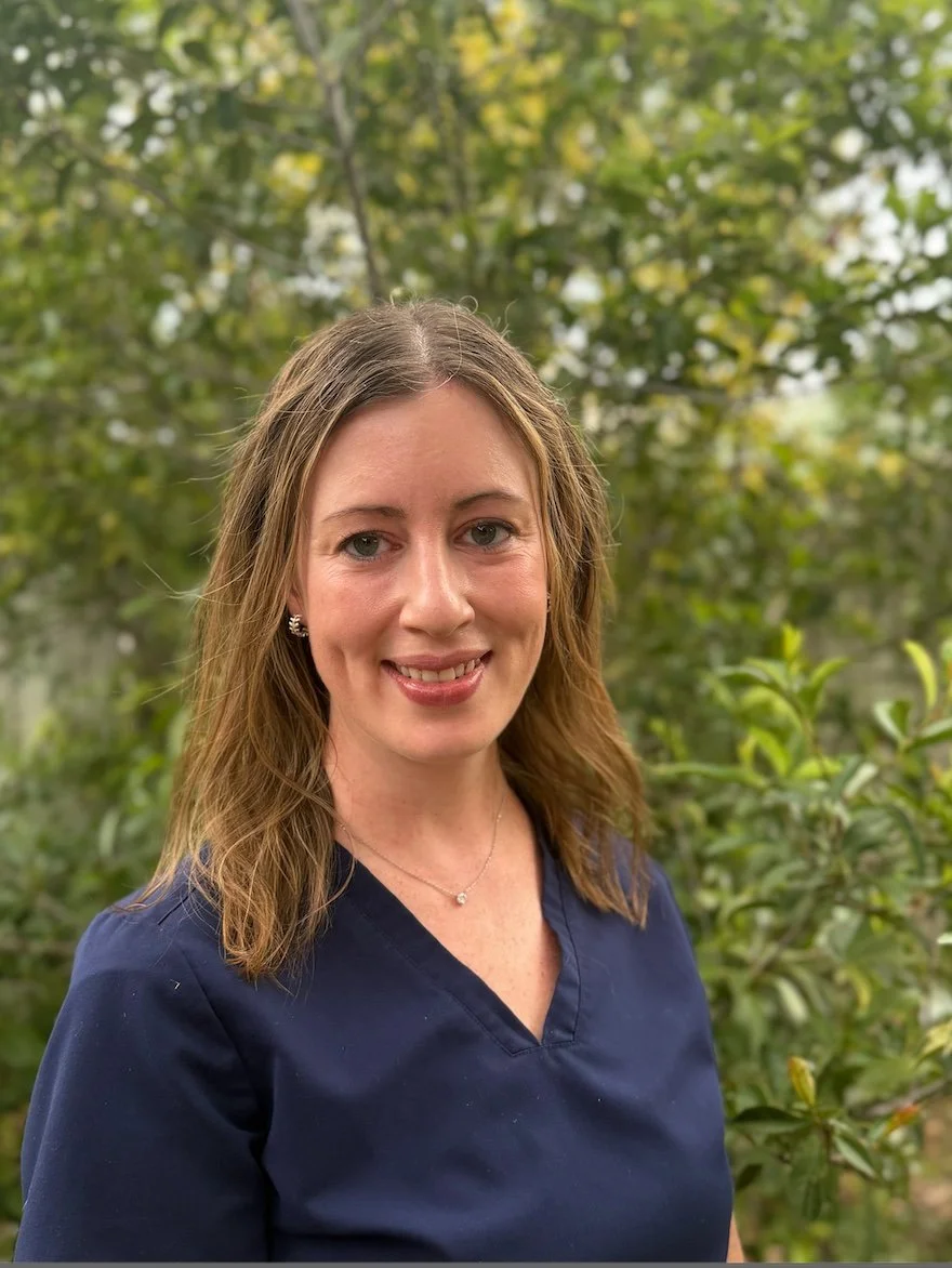 A woman with shoulder-length brown hair, wearing a navy blue top and a small necklace, standing outdoors with green trees in the background.