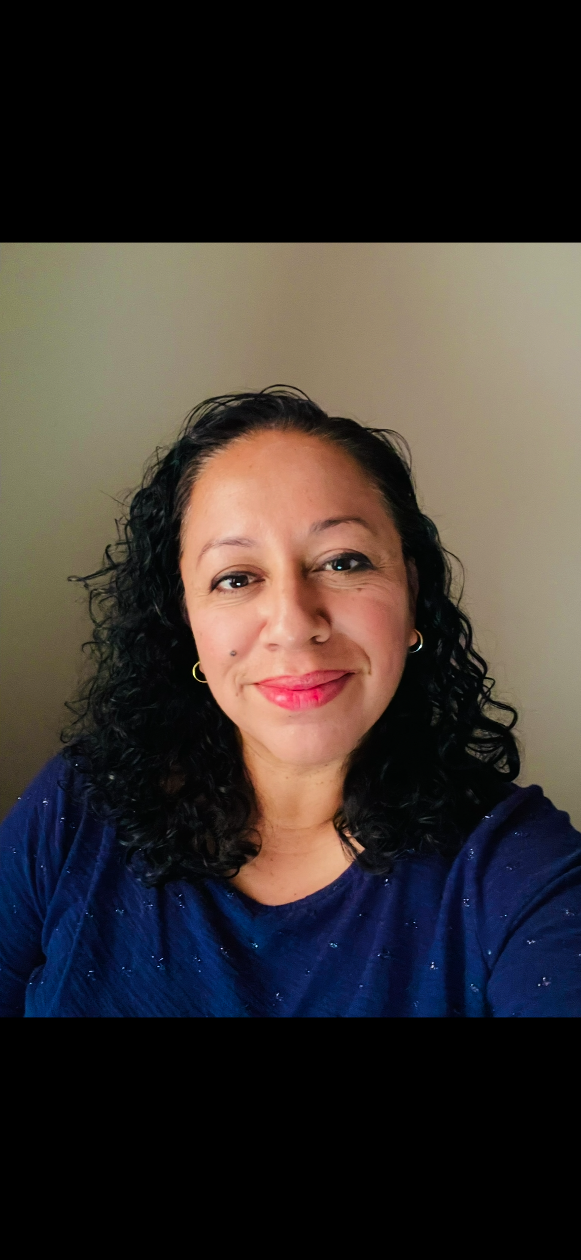 A woman with curly black hair wearing a navy blue shirt and gold hoop earrings, smiling against a plain beige wall.