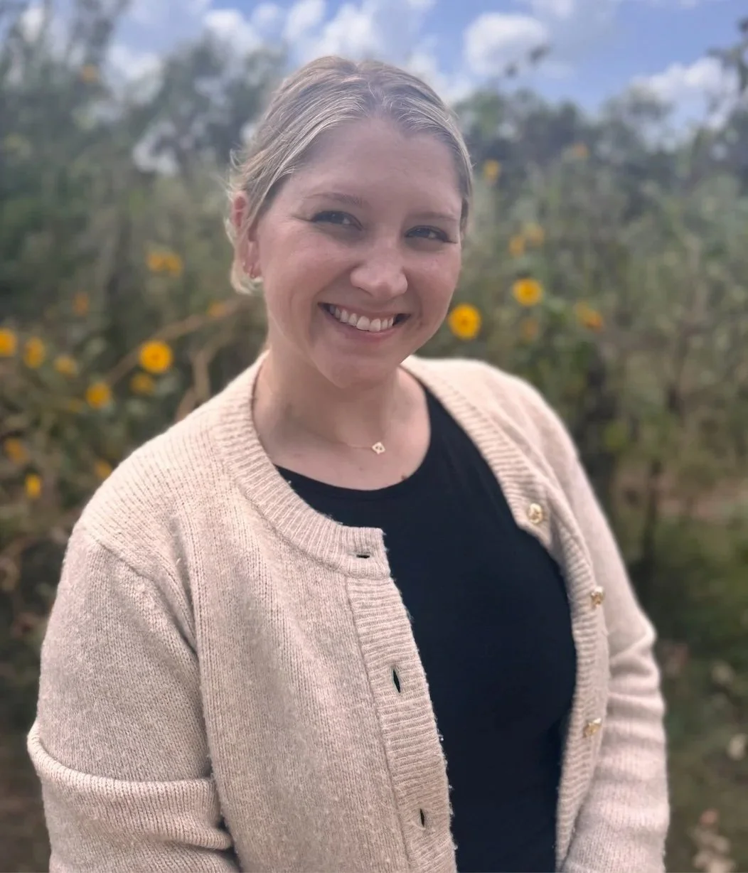 A smiling woman with blonde hair in a low bun, wearing a beige cardigan over a black top, standing outdoors with yellow flowers and green foliage in the background.