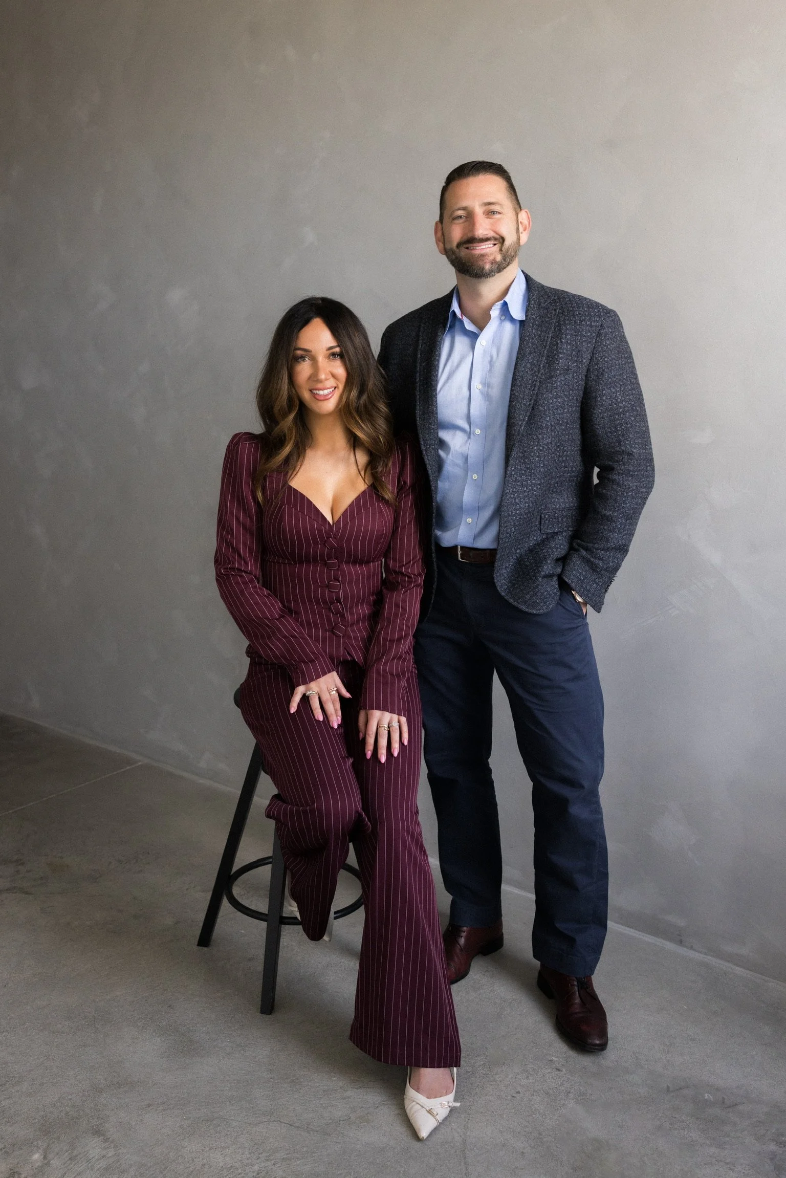 A man and woman posing together indoors against a plain gray wall, smiling at the camera. The woman is seated on a black stool, wearing a burgundy pinstripe suit with wide-leg pants, and beige high heels. The man stands next to her, dressed in a blue shirt, dark gray patterned blazer, navy pants, and brown shoes.