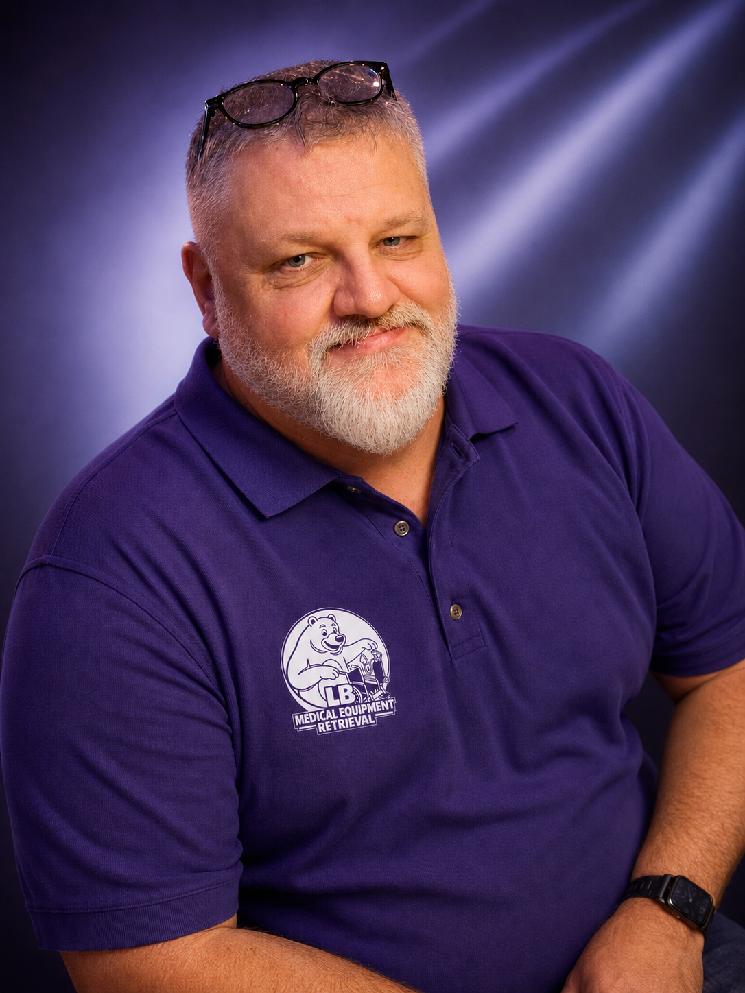 A middle-aged man with glasses on his head and a beard, wearing a purple polo shirt with a logo of a bear holding tools and the text 'Medical Equipment Retrieval' on it, smiling and sitting against a dark, blurred background.