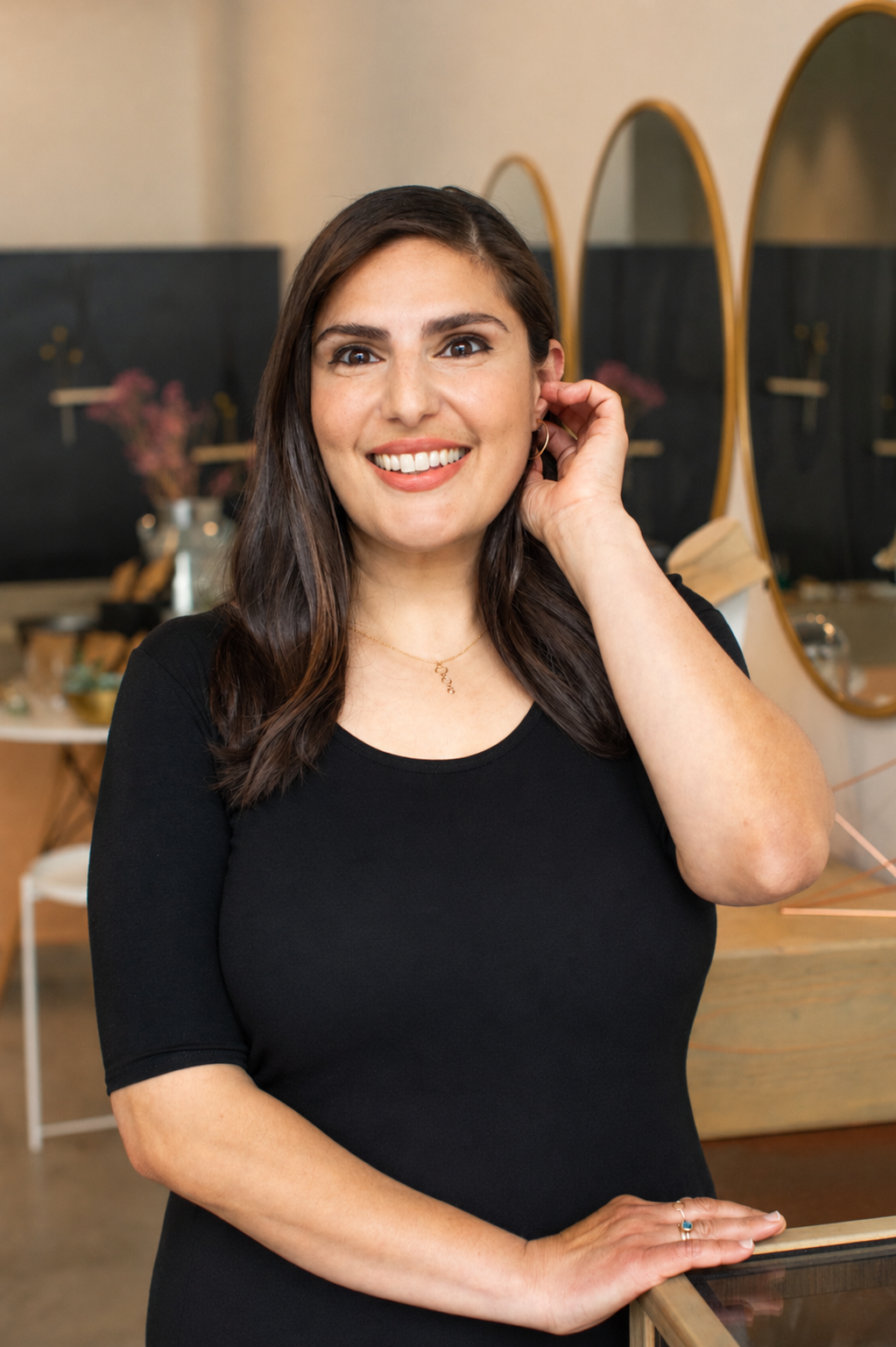 A woman in a black shirt smiling, touching her ear, with mirrors and a display of flowers in the background.