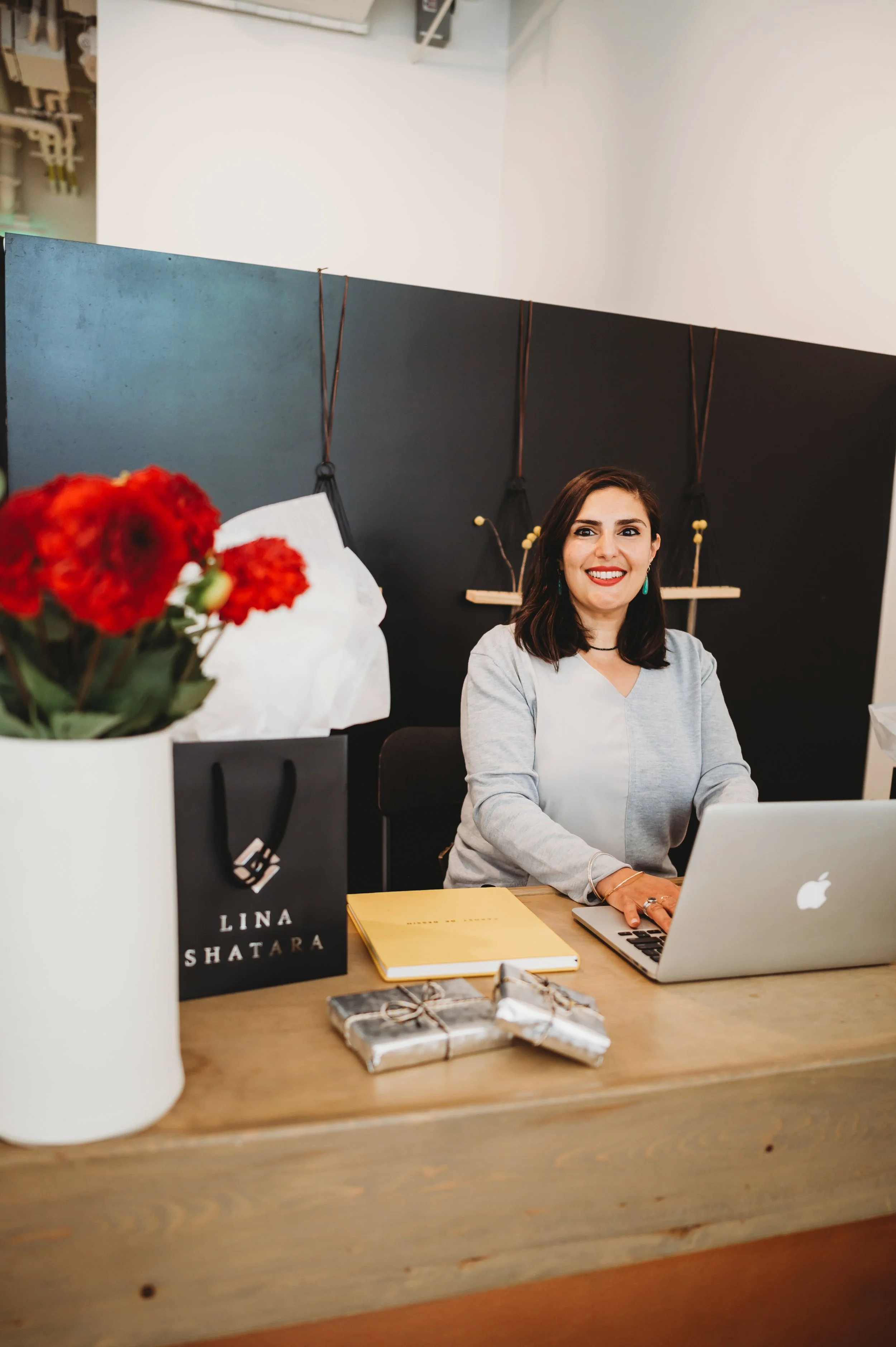 A woman with dark hair and earrings smiling while working on a laptop at a wooden desk. The background features a modern, stylish wall with hooks and decorative elements. On the desk, there are a yellow notebook, small wrapped gifts, a flower bouquet, and a black paper bag with the name 'LINA SHATARA' printed on it.