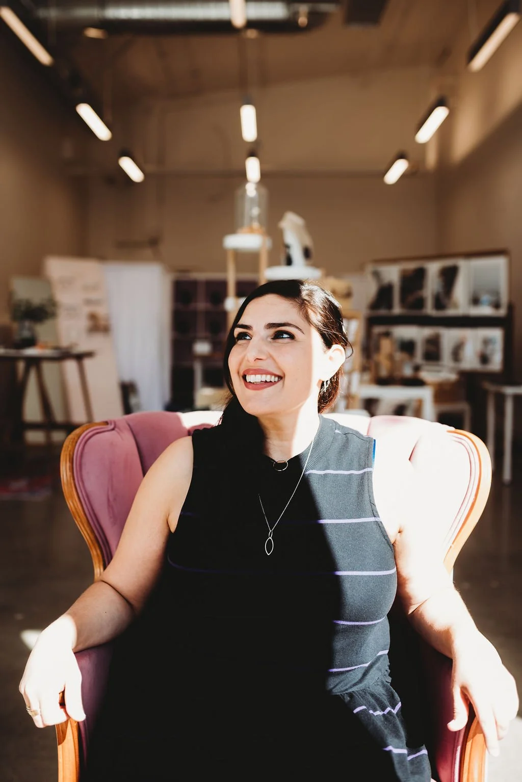 A young woman with dark hair sitting on a pink upholstered chair, smiling and looking to her left, in a well-lit room with artistic and decorative items in the background.