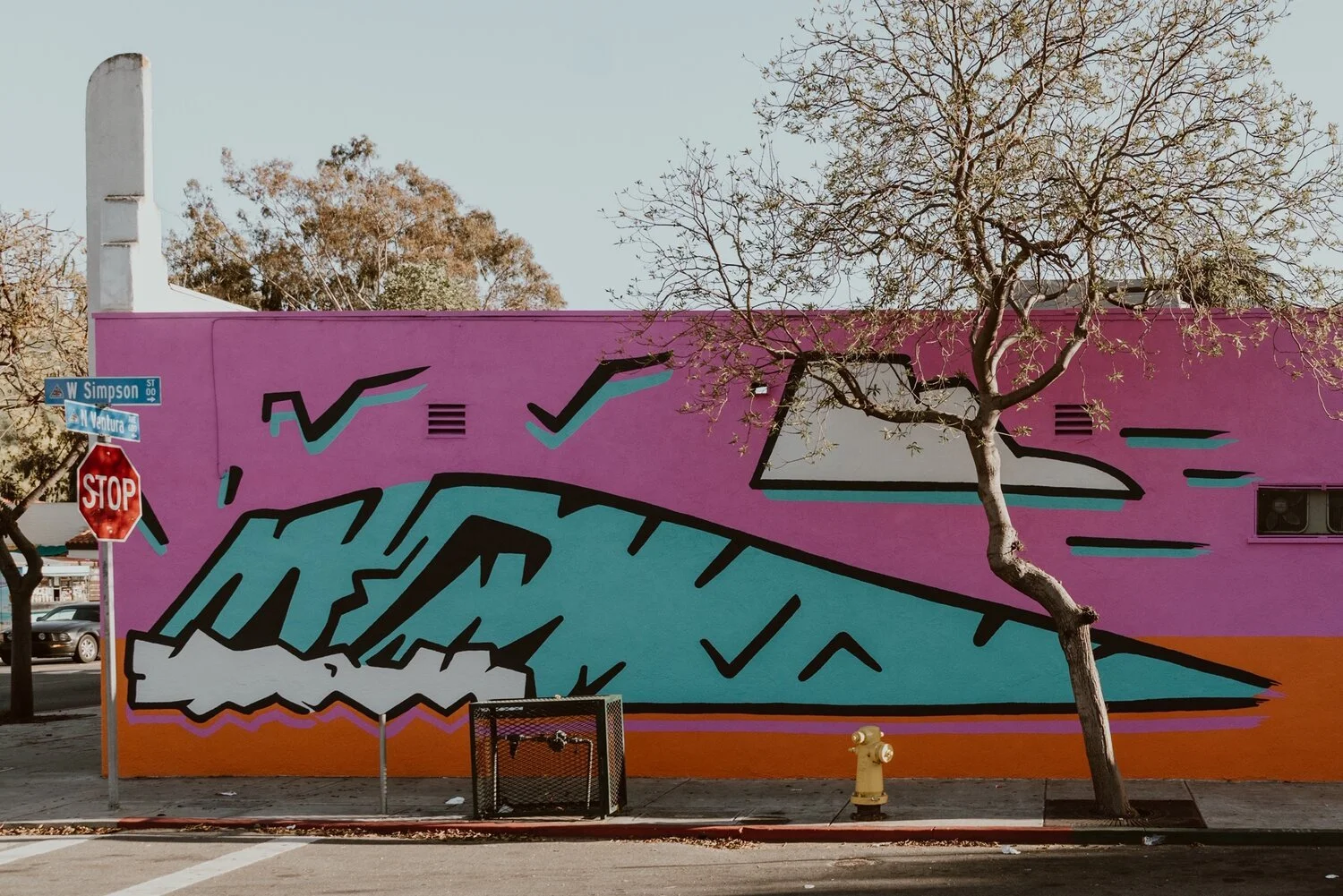 Colorful street mural with a large stylized wave, geometric shapes, and check marks, painted on a wall behind a leafless tree in an urban setting.