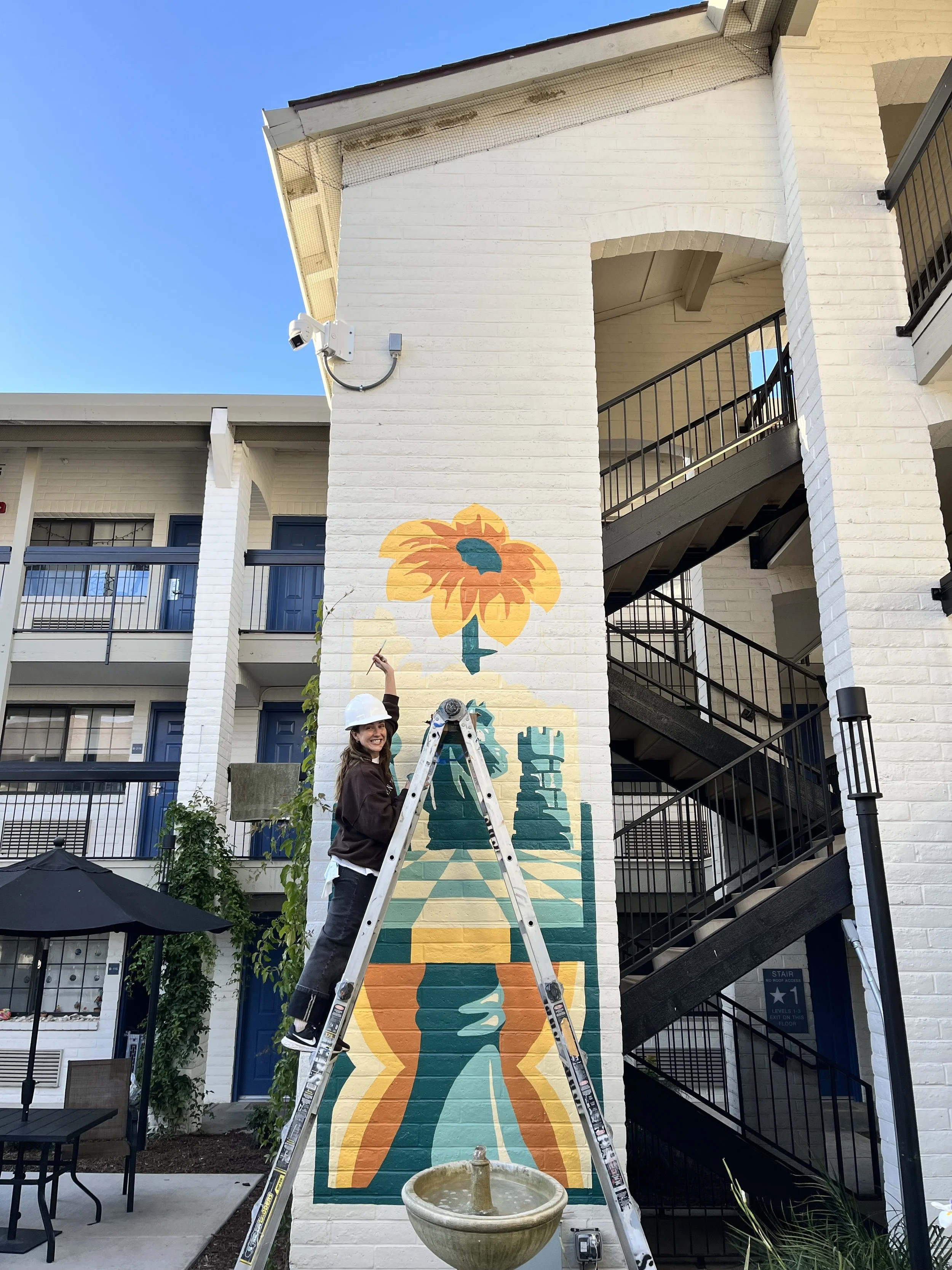 A woman wearing a white hard hat and dark jacket is standing on a ladder, painting a mural of the Statue of Liberty on a white brick wall of a building. The mural features a simplified illustration of the statue with an orange and teal color scheme. The scene is outdoors during daytime, with a blue sky and adjacent building with balconies visible in the background.