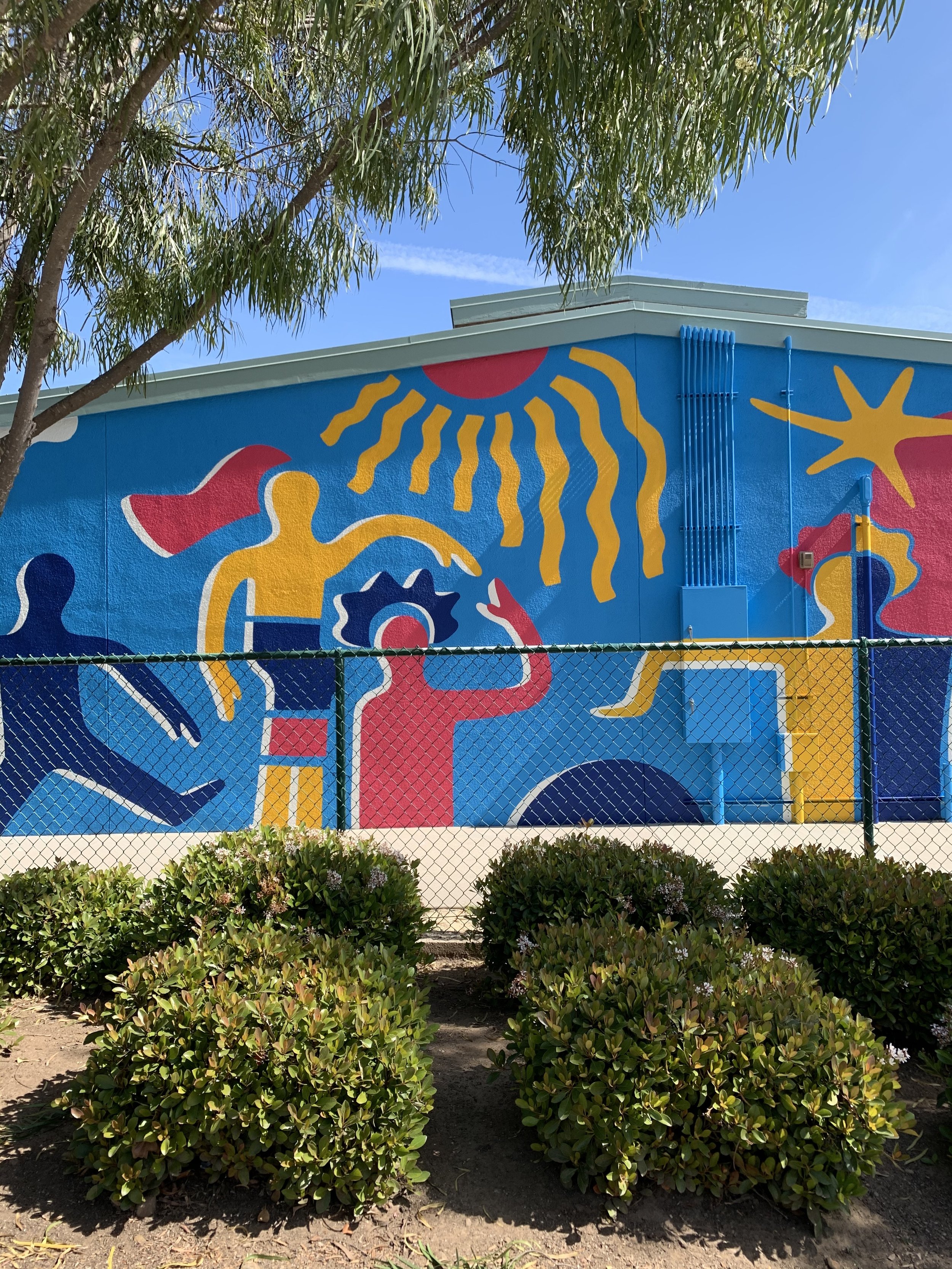 Colorful painted mural on a wall behind a chain-link fence, with green bushes in foreground and a tree with leaves partially obscuring the top of the mural, under a blue sky.