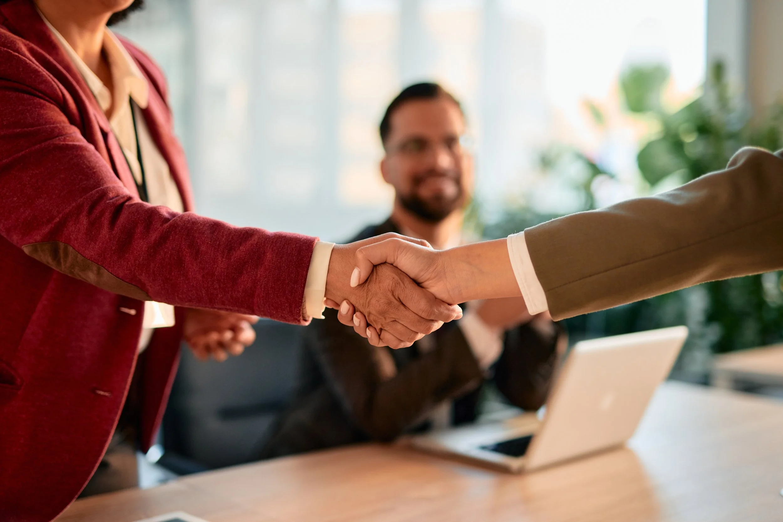 Two business women shaking hands. Close up of the hand shake with a man blurred behind looking on.