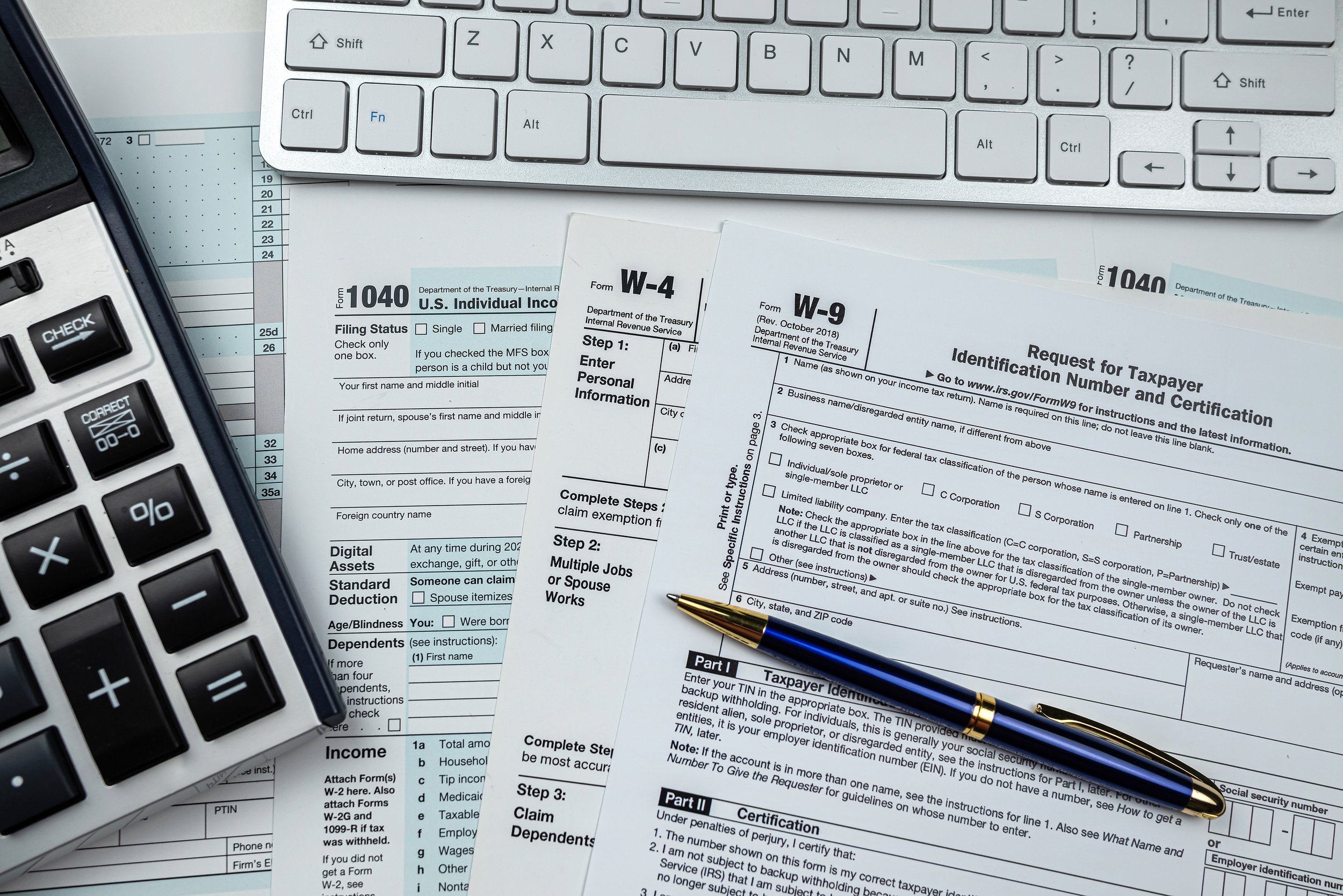 Various tax forms such as 1040, W-4 and W-9 spread across a desk with a keyboard above them and blue pen on top of them.