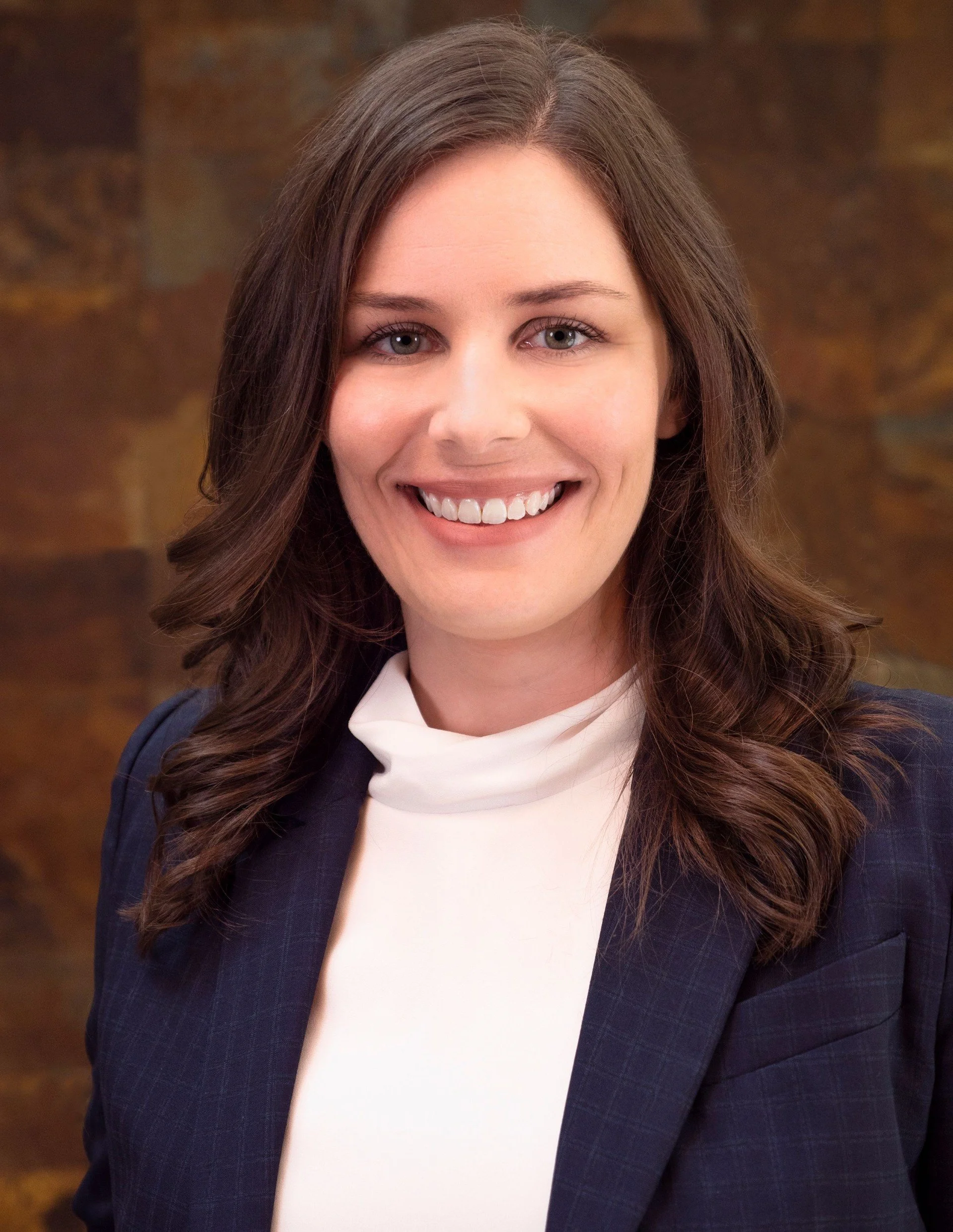 Close-up of a woman with long brown hair and gray eyes, smiling, wearing a white turtleneck and a navy blazer with a subtle checkered pattern, standing in front of a blurred brick wall background.