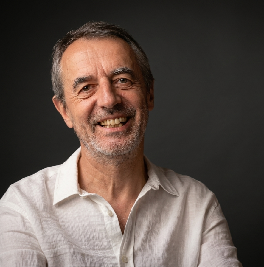 Hombre mayor de cabello gris y barba, sonriendo, usando camisa blanca, fondo oscuro.